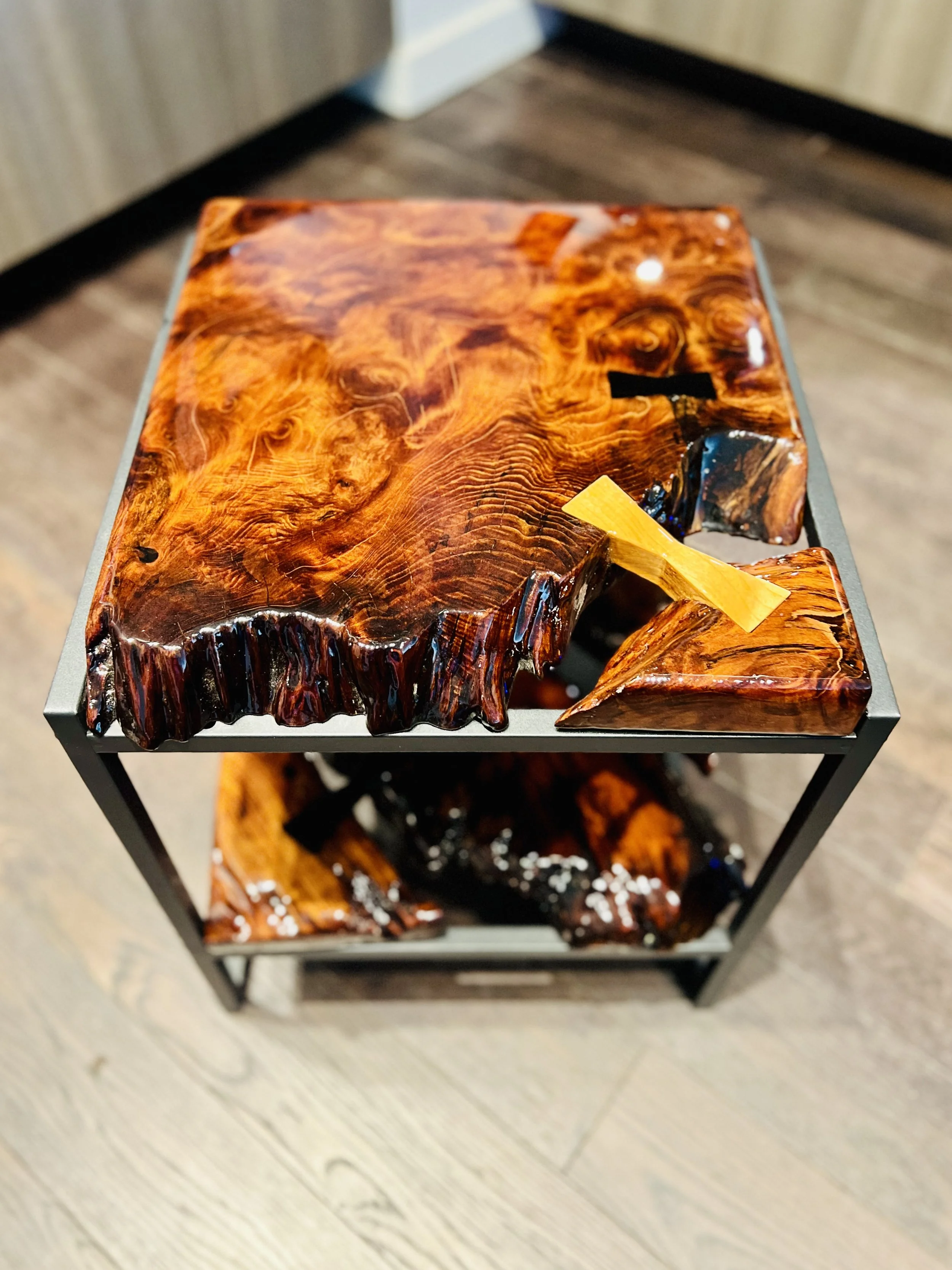 A wooden side table with a polished, knotty, and glossy wood surface, showcasing rich, swirling wood grain patterns. The table has a black metal frame and a lower shelf, also made of polished wood.
