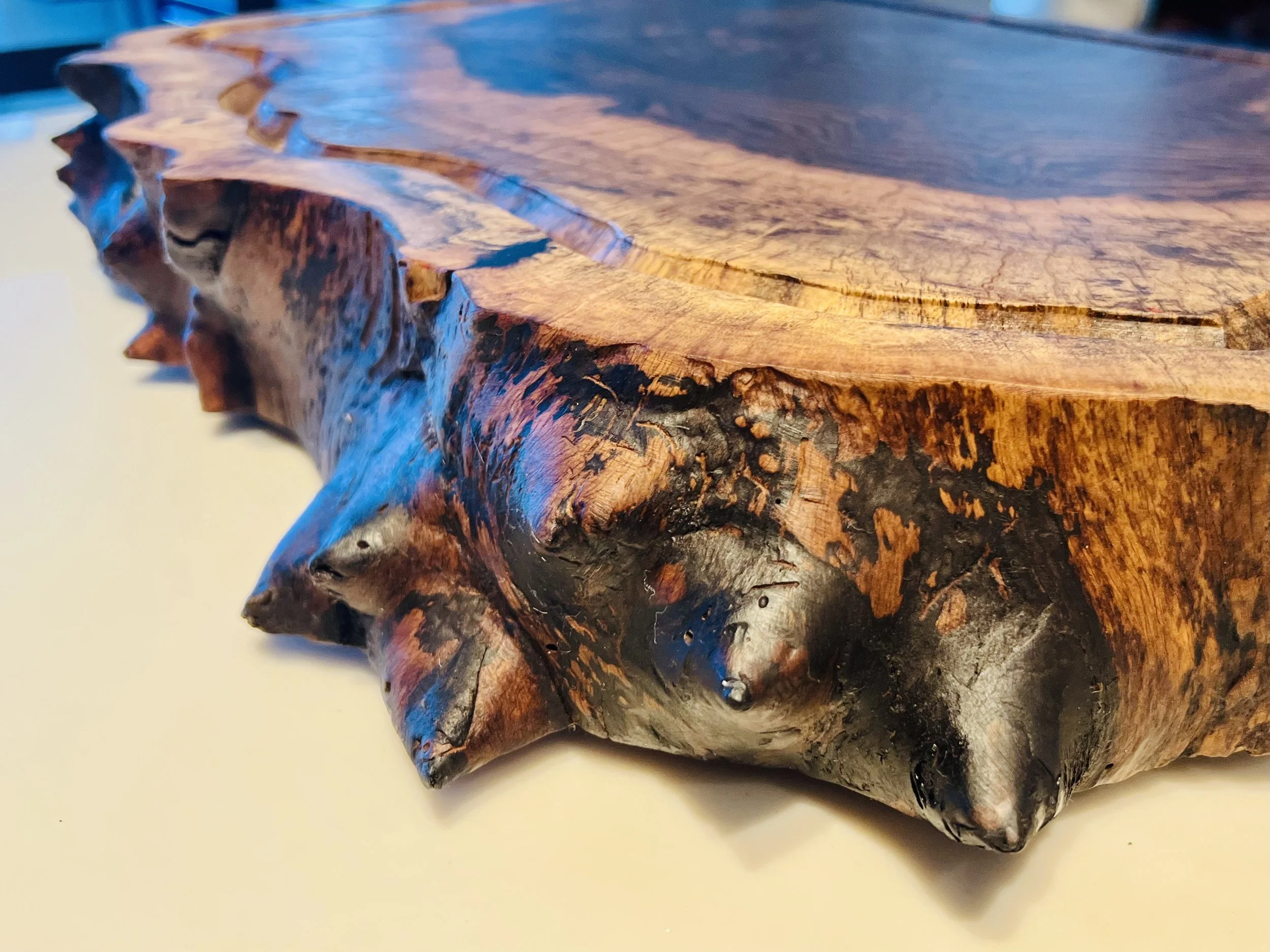 Close-up view of a polished, irregularly shaped wooden table or slab with a natural edge, showing wood grain and textured bark.