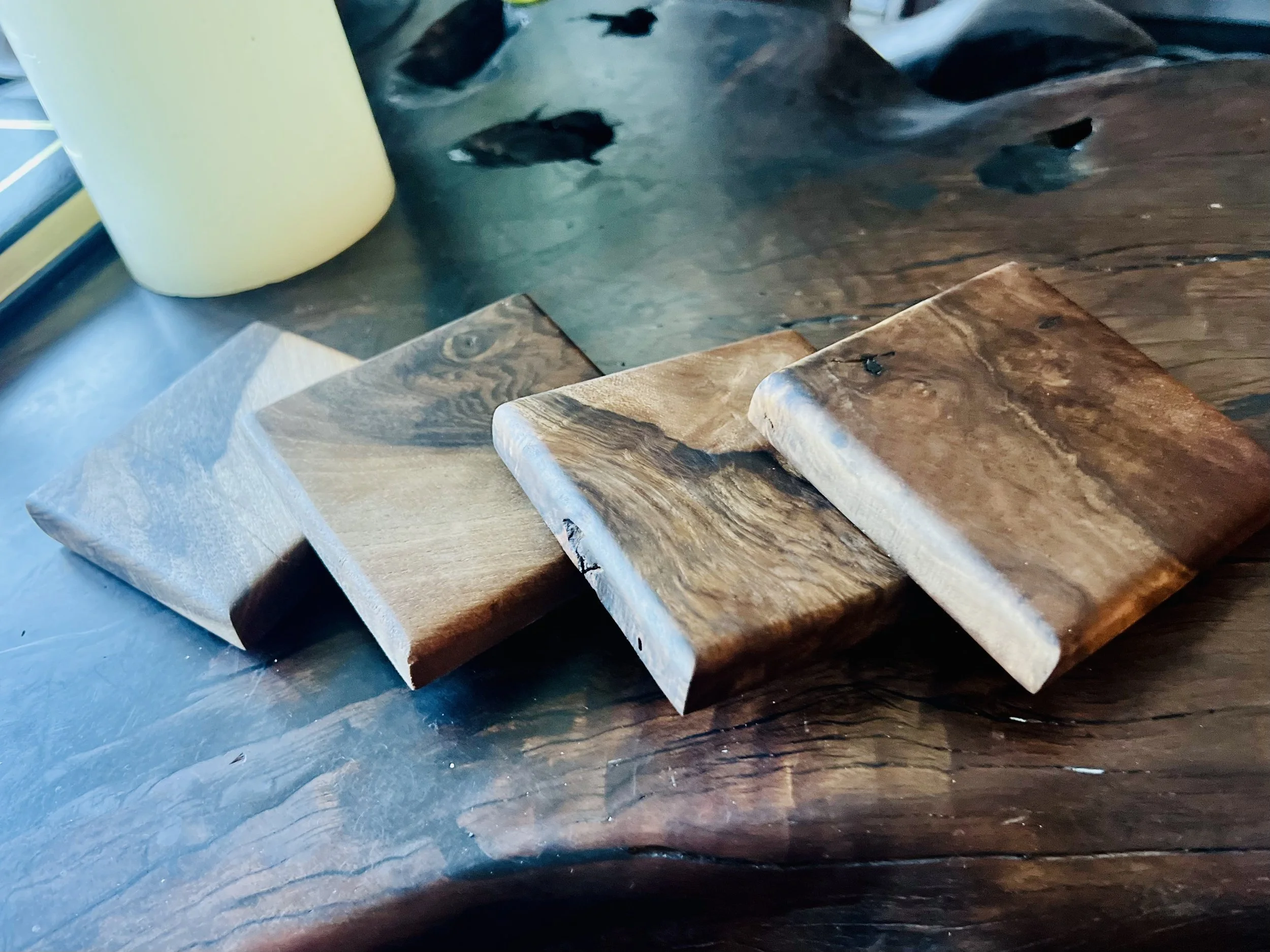 Five different sold walnut wood coasters placed on a dark wooden table near a window, with a yellow container in the background.