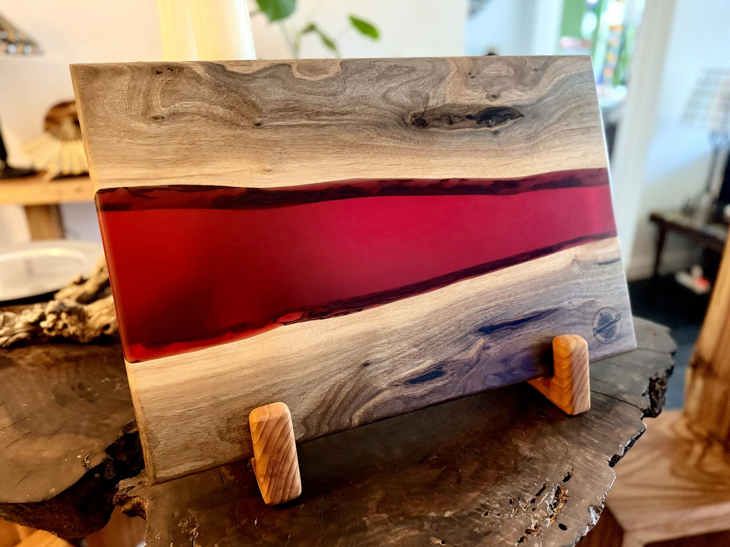 A wooden and resin art piece displayed on a small wooden stand, with a natural wood grain pattern and a prominent horizontal red resin stripe.