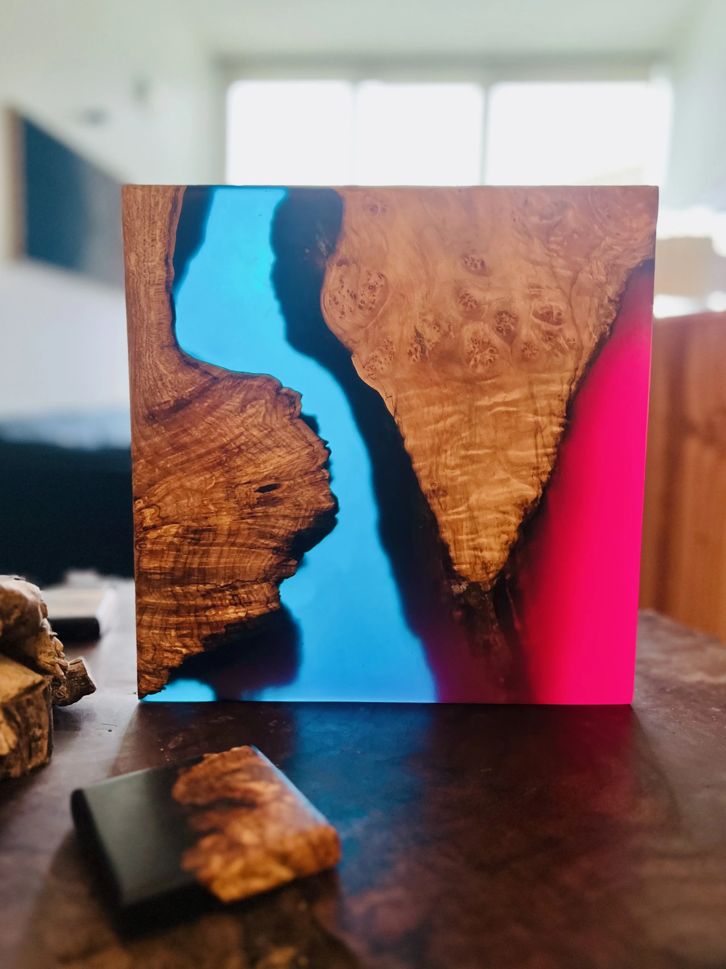 A wooden art piece with a blue resin river running through the middle, placed on a wooden table with some smaller wood pieces nearby, in a bright room with windows in the background.