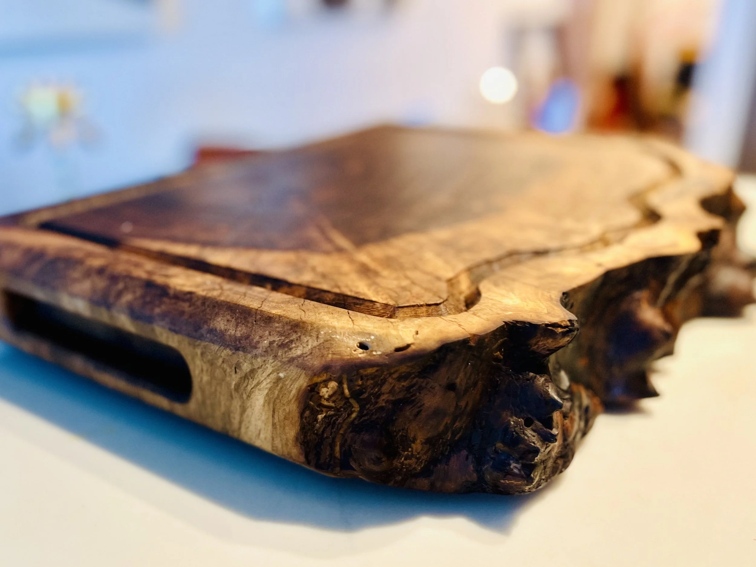 Close-up of a rustic, handcrafted wooden serving or cutting tray with natural edges, placed on a white surface, with a blurred background.