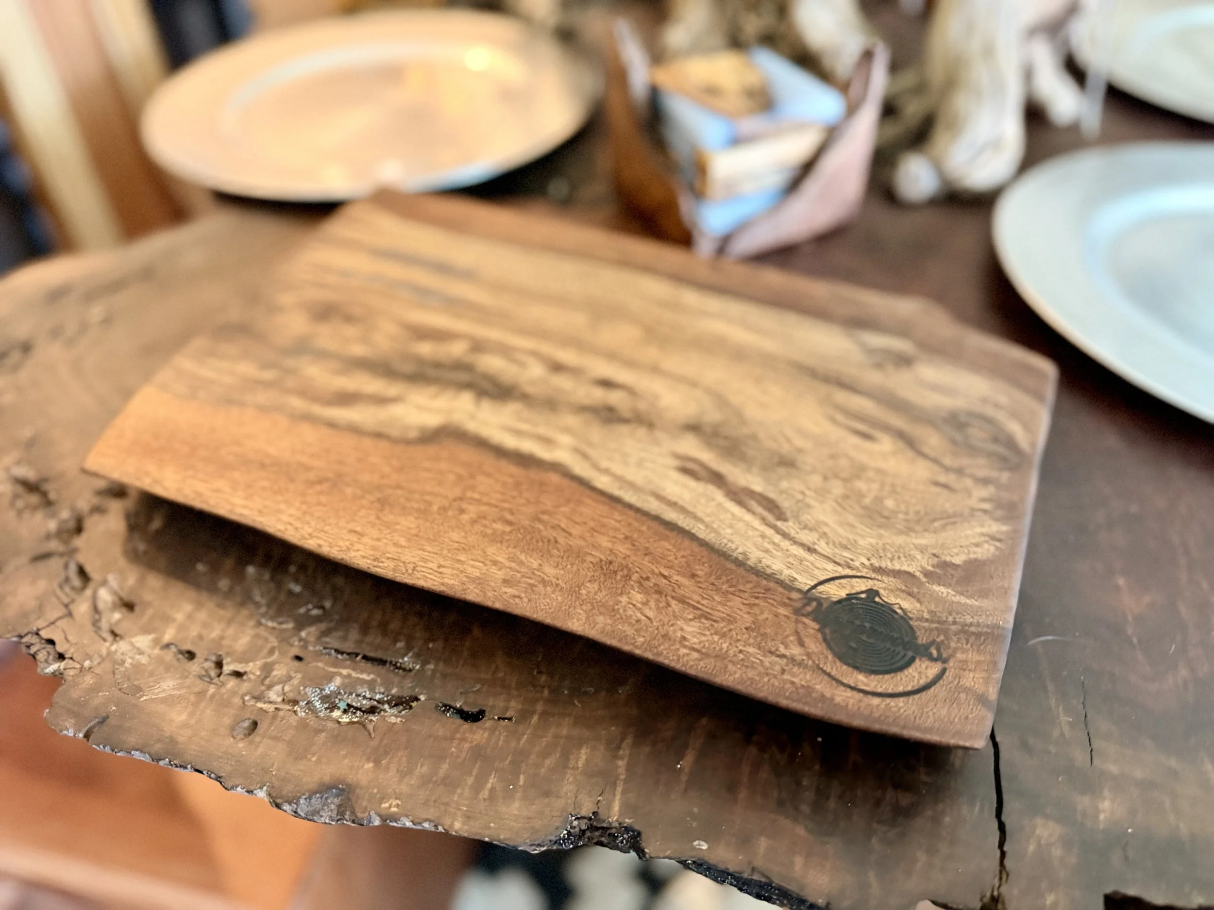 Close-up of a wooden cutting board with a natural finish and a small black engraved insect design on one corner, placed on a rustic wooden surface.