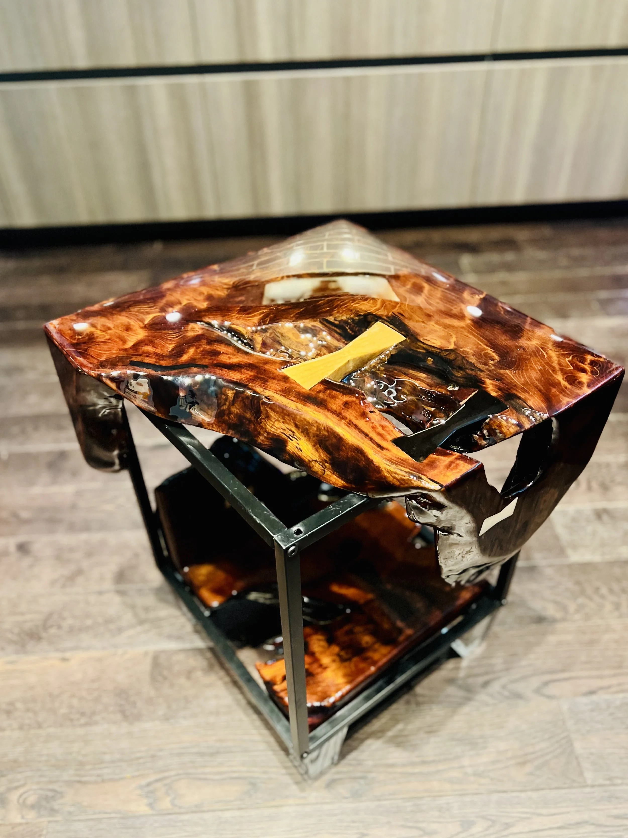 A small, elevated table with a glossy, wavy, dark brown and black surface, resembling animal wood grain, with a yellow and black object on top, set on a wooden floor with a beige wall in the background.