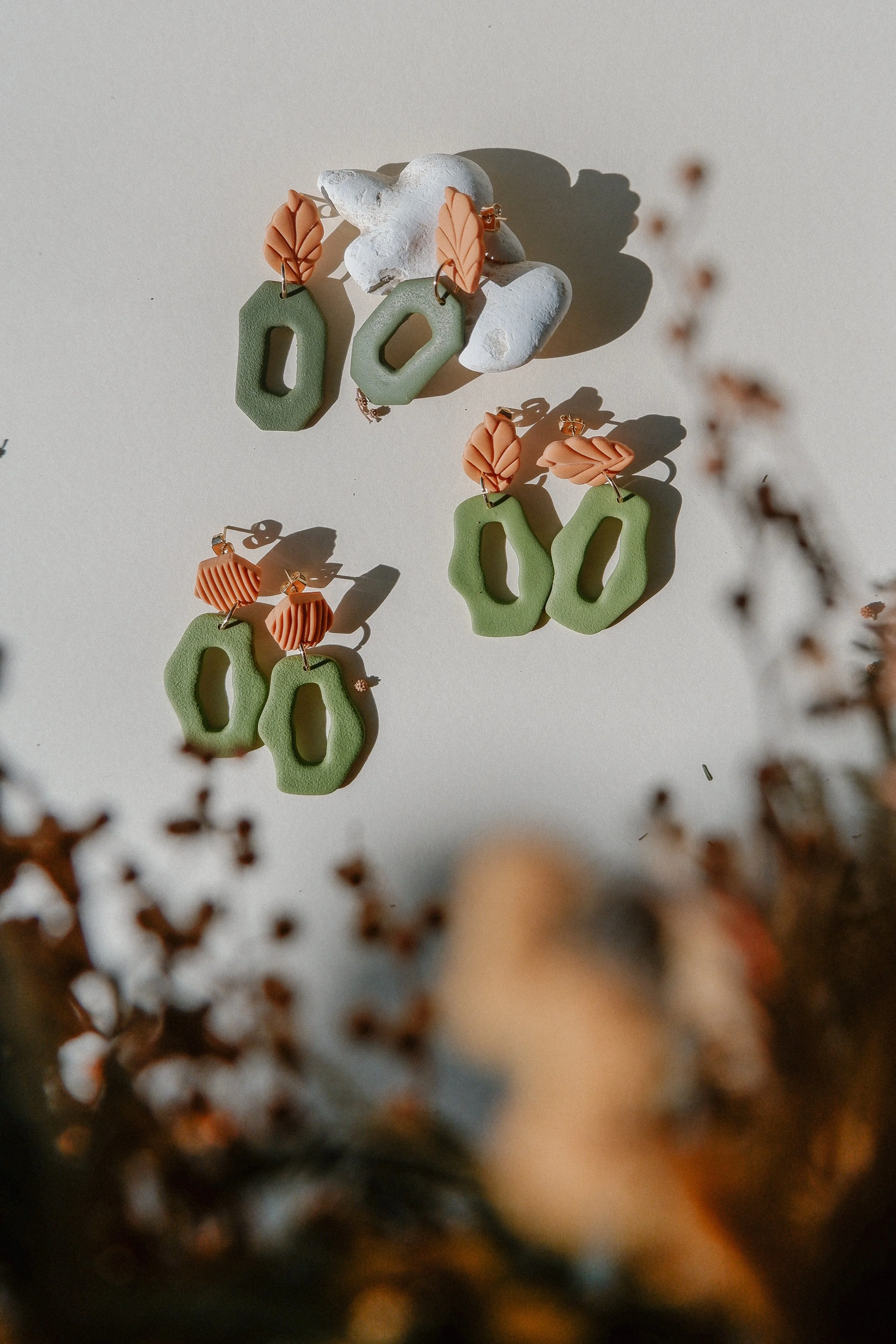 Colorful, geometric earrings with leaf-shaped accents, displayed on a white background with decorative branches in the foreground.