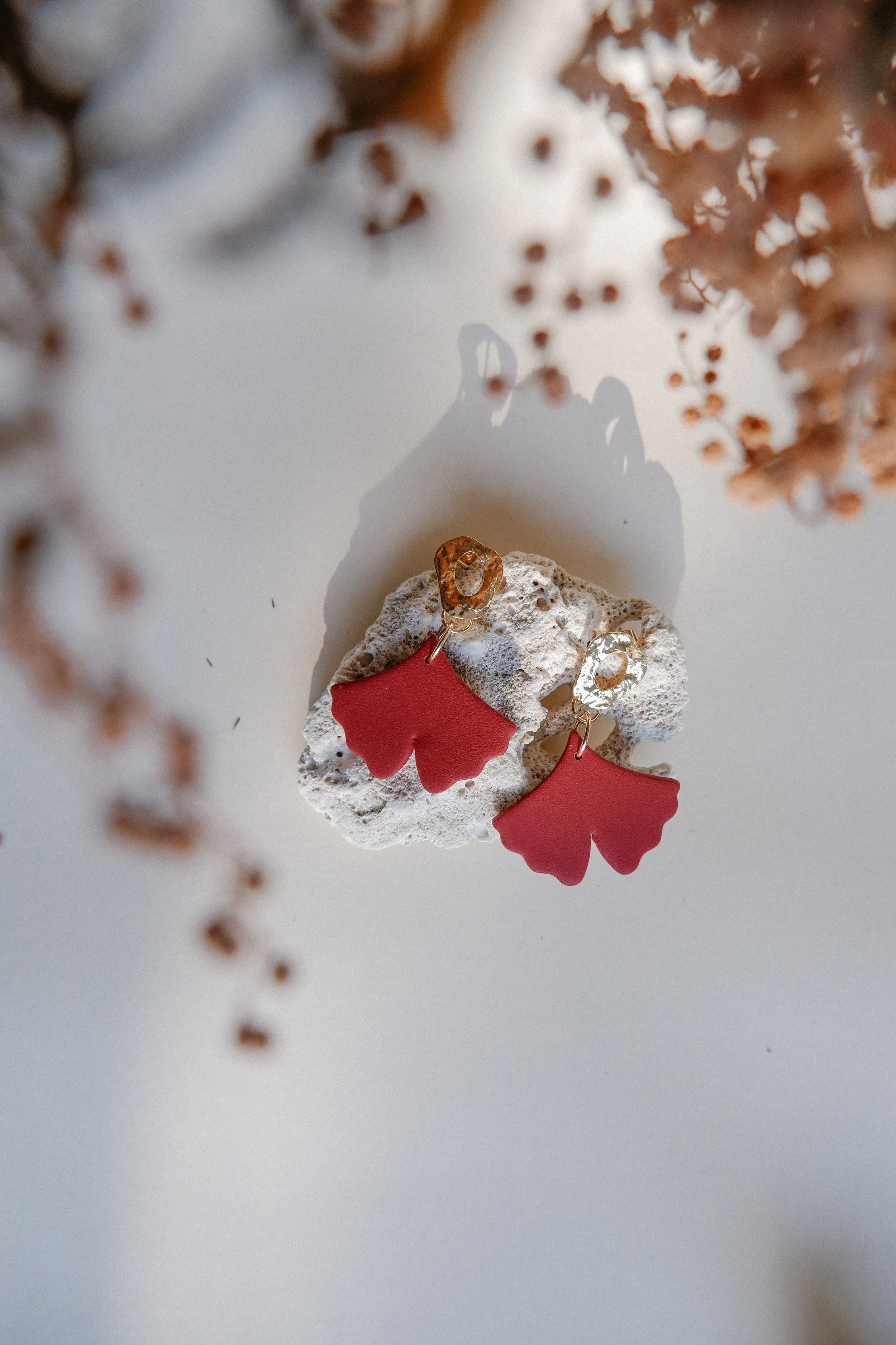 Pink flower-shaped earrings with gold hooks resting on a piece of coral, with dried flowers and shadows in the background.