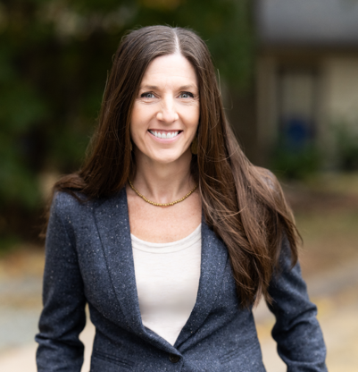 A woman with long brown hair, blue eyes, and a bright smile standing outdoors in front of a blurred background of trees and a brick building.