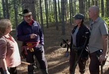 Four people stand talking in a sunlit pine forest. A man in a black cowboy hat gestures while the others listen.