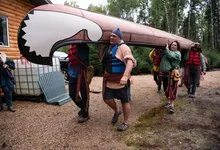 A group carries a large canoe with a stylized animal-head design overhead along a gravel path beside a log building.
