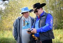 Two men, one wearing a blue bucket hat and the other a black cowboy hat, examine a handheld device beside autumn woodland.