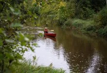 A paddler in a red canoe travels along a narrow, calm river framed by leafy branches and forest.
