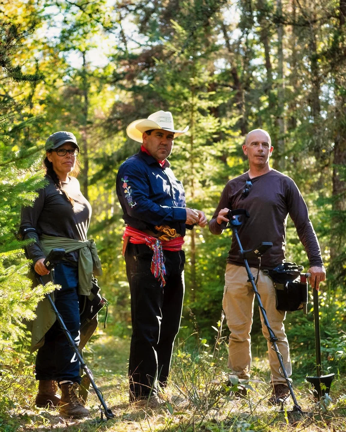 Laurie (Goldie), Memphis and Eric, the gold-hunting team from Red River Gold Season 2, stand in a sunlit forest with metal detectors and digging equipment. Memphis wears a white cowboy hat and a colourful woven sash.