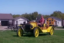 A man sits at the wheel of a yellow antique open-top car parked on grass beside historic buildings and farm equipment.