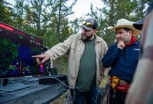 A man points to a colourful sonar display while another man in a cowboy hat studies the screen aboard a boat.