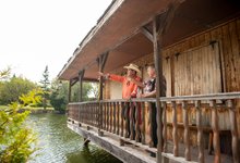Two people stand on the covered wooden balcony of a rustic waterfront building, one pointing across the water.