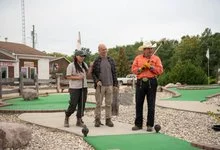 Three people stand on a miniature golf course. A woman lines up her putt while two men watch.