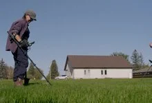 A woman wearing a cap and backpack sweeps a metal detector over grass near a small white church.