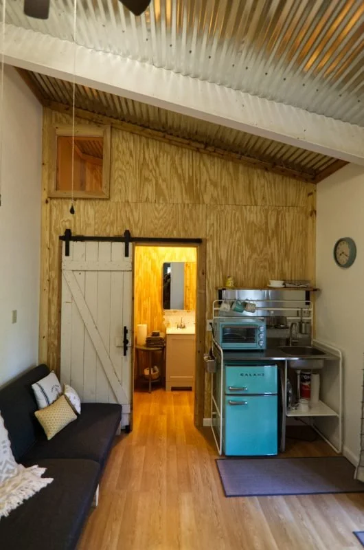 Interior view of a cozy room with a rustic wooden wall and ceiling, a black sofa with decorative pillows, a blue mini-fridge, a small microwave, and a kitchen counter, with a doorway leading to a bathroom