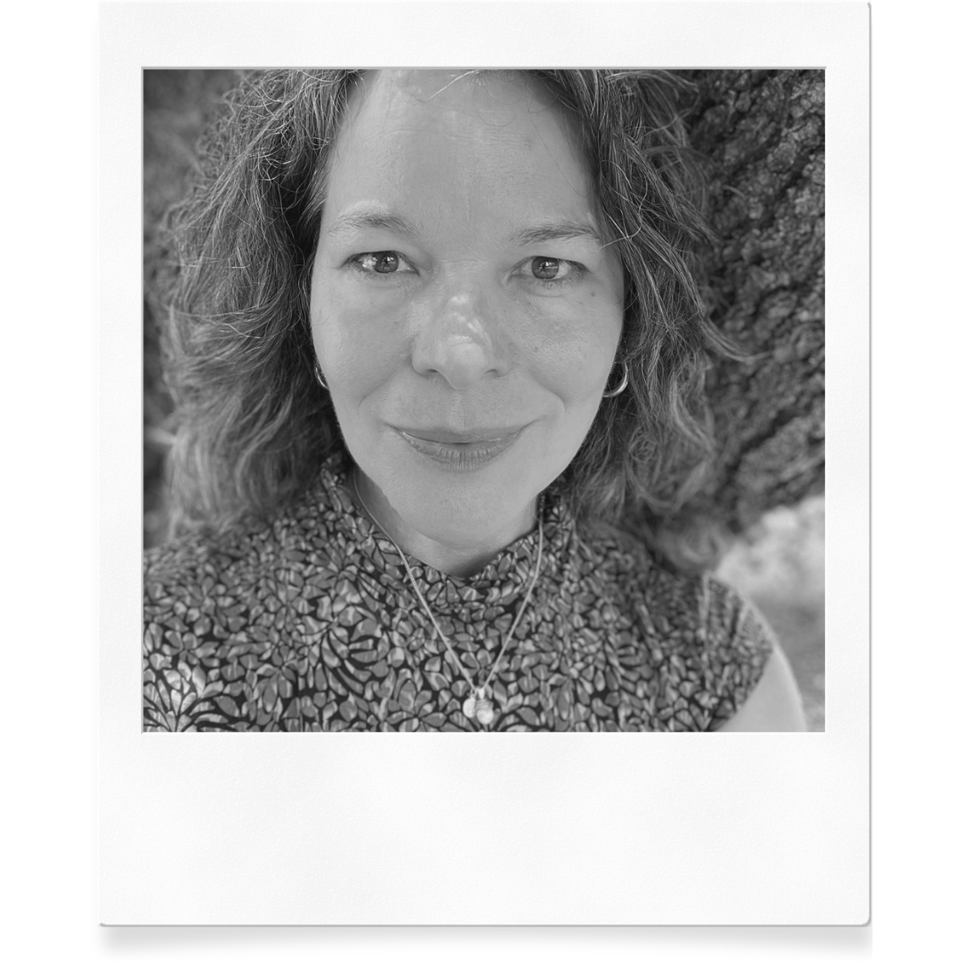 Black and white close-up portrait of a woman with wavy hair, wearing a patterned top, earrings, and a necklace with a pendant, outdoors with trees in the background.