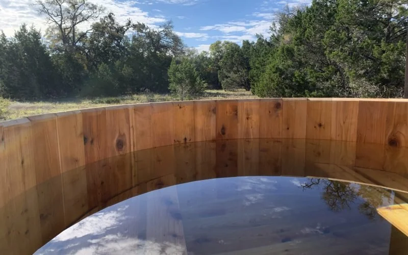 A wooden hot tub filled with water outdoors, with a view of trees and a cloudy sky in the background.