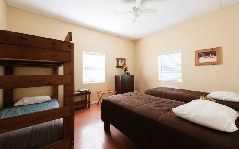 A bedroom with three beds, two single beds and a bunk bed, in a room with beige walls, windows with blinds, a ceiling fan, a wooden dresser with a flower vase, and a framed picture on the wall.