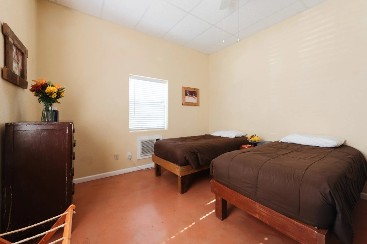 A bedroom with two beds covered in brown comforters and white pillows, a window with blinds, a small framed picture on the wall, a dresser with a vase of flowers, and a small nightstand between the beds with flowers and a book.
