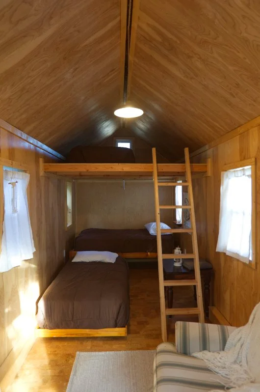 A cozy wooden cabin room with two beds, one on the floor and a bunk bed, light coming through small windows, ladder leading to upper bunk, and a striped armchair in the corner.