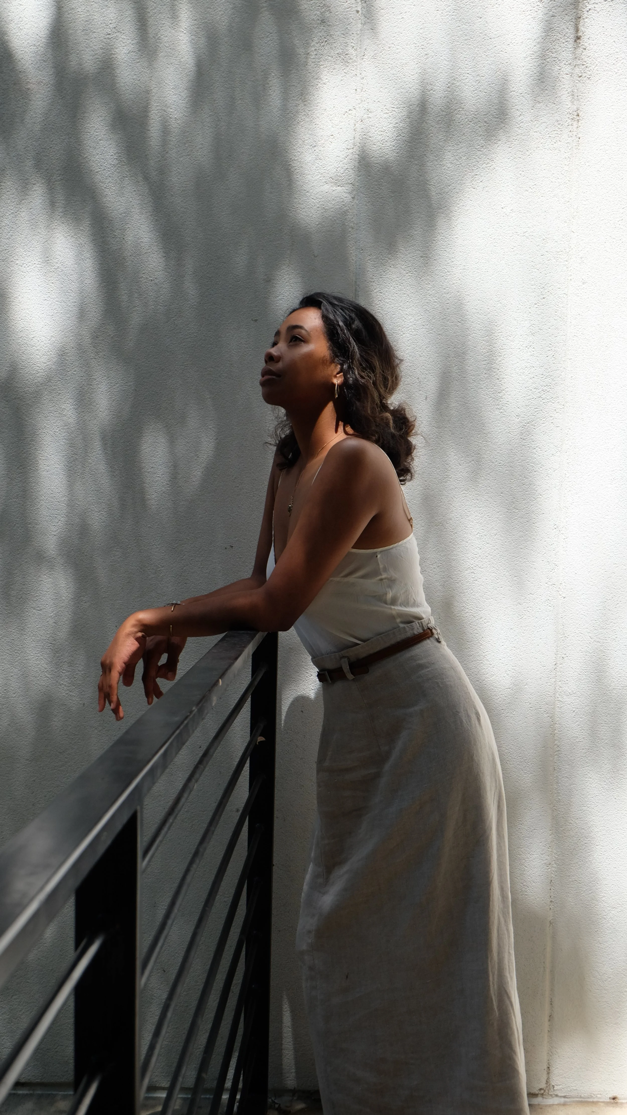 A woman leaning on a railing outside, with shadows of tree leaves cast on a white wall behind her.