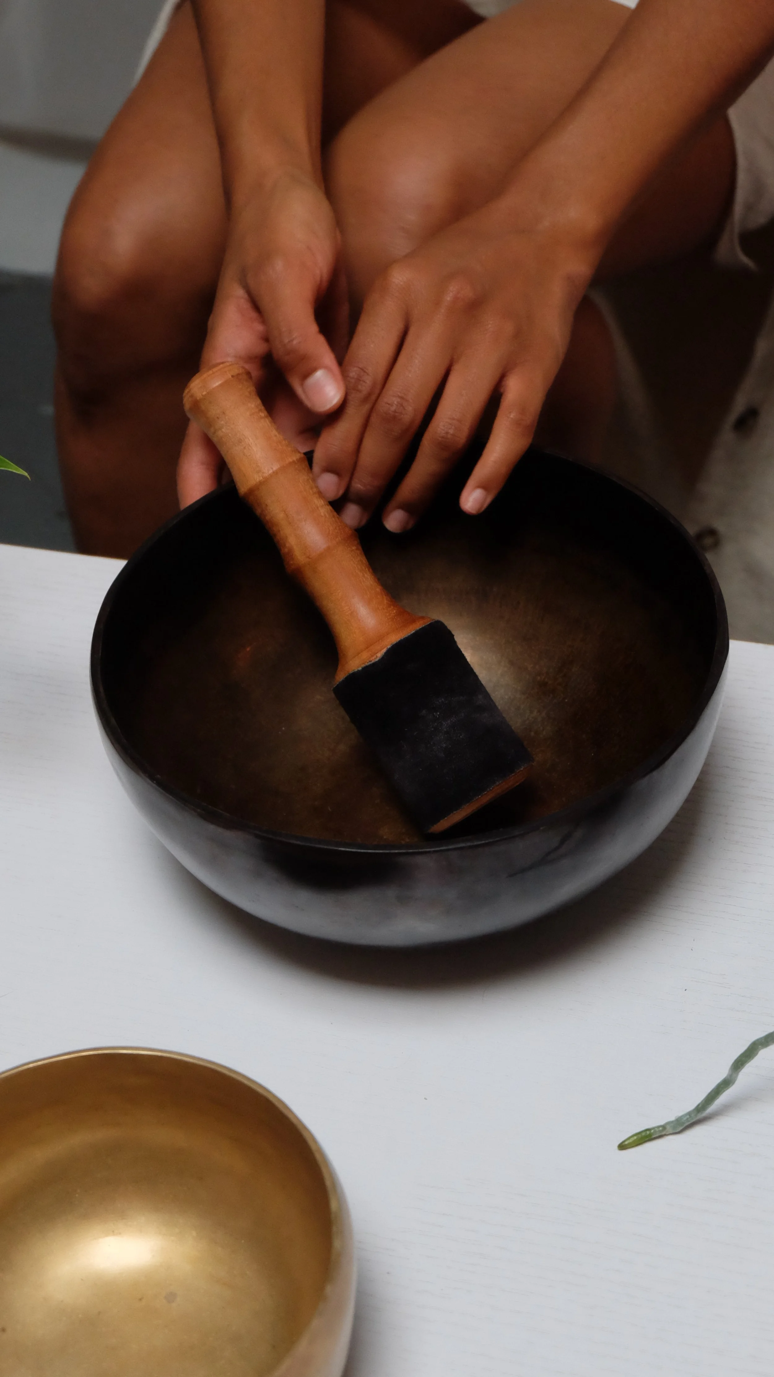 Person holding a wooden hammer over a black singing bowl, another gold-colored singing bowl on a white table.