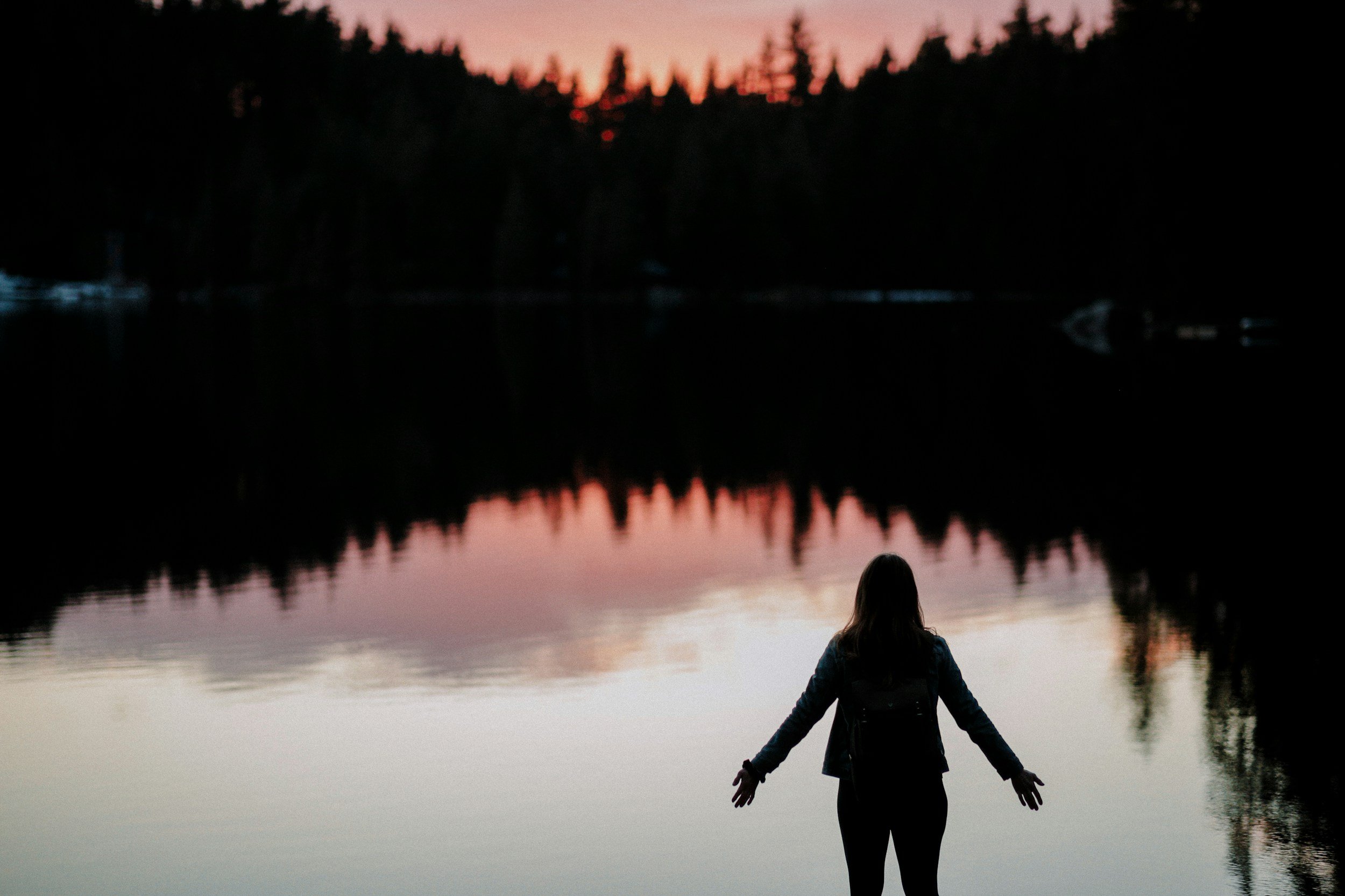 A person standing at the edge of a calm lake during sunset, facing the water with outstretched arms, surrounded by silhouetted trees and a pink-orange sky reflected on the water, representing a Christian woman healing with EMDR and OCD therapy.
