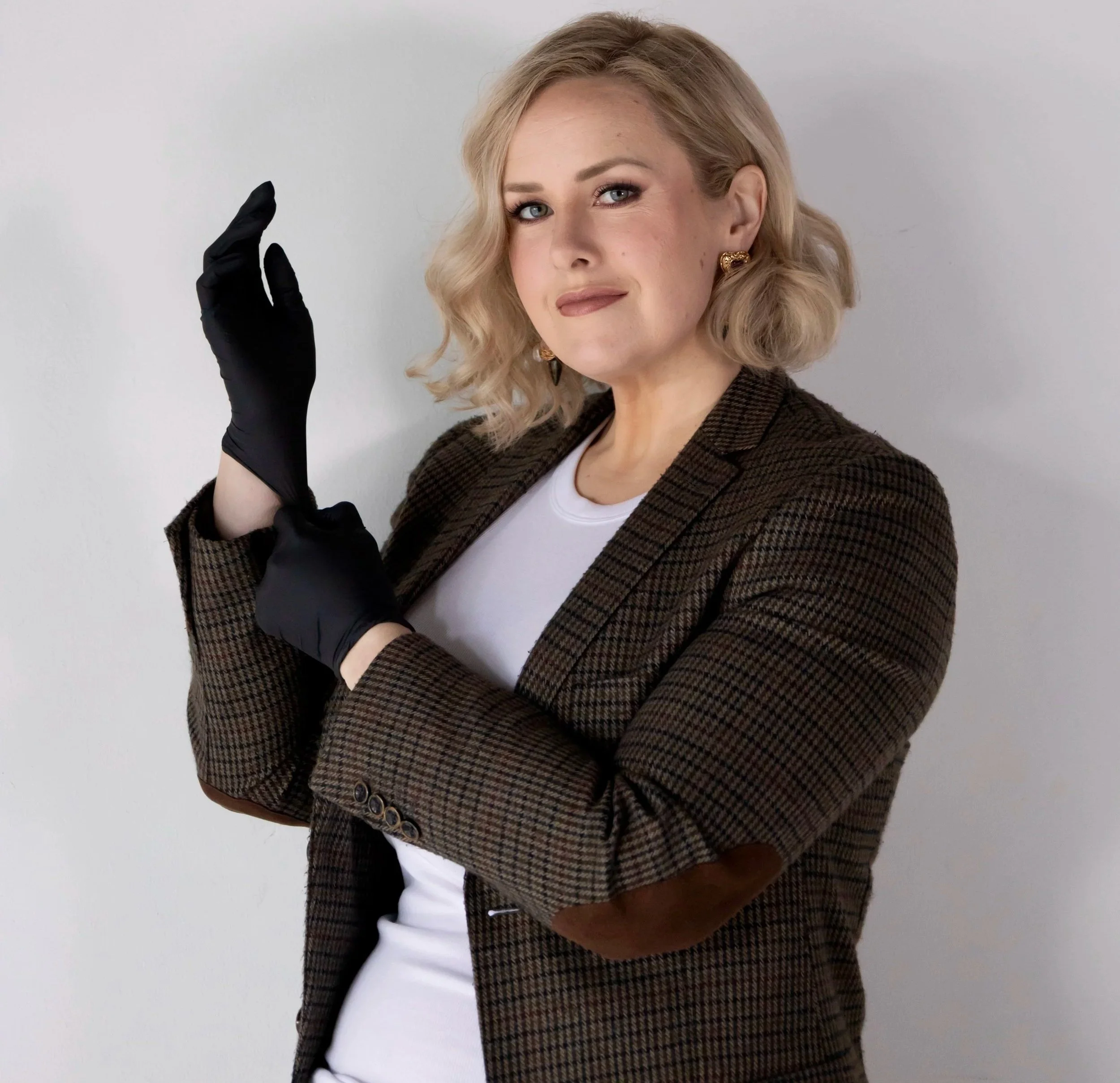 A woman with blonde, wavy hair smiling outdoors with greenery in the background.