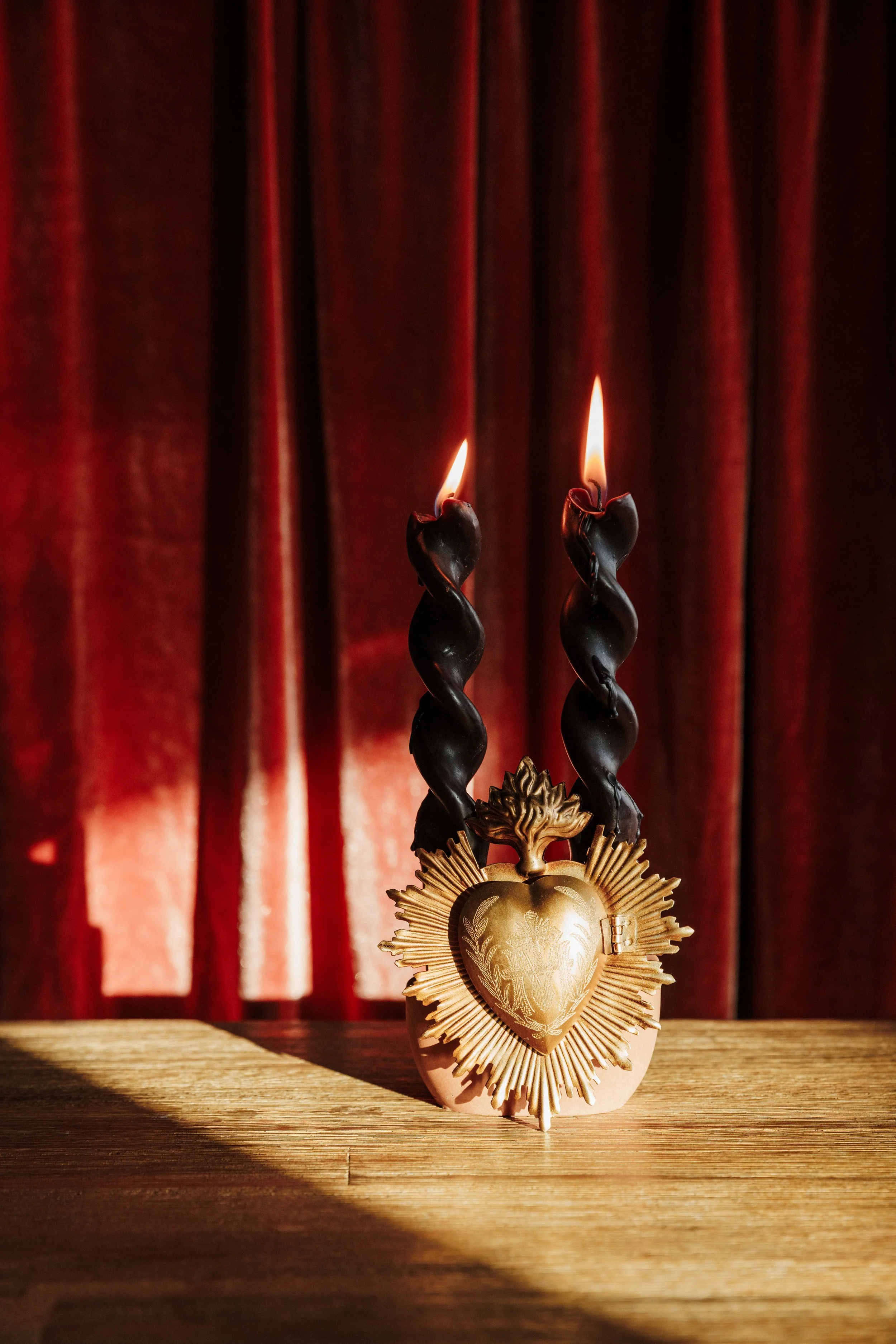 An ornate religious medal with a heart shape and a crown of thorns, suspended above a wooden surface. Two twisted black candles with flames are behind the medal, against a backdrop of red curtains.