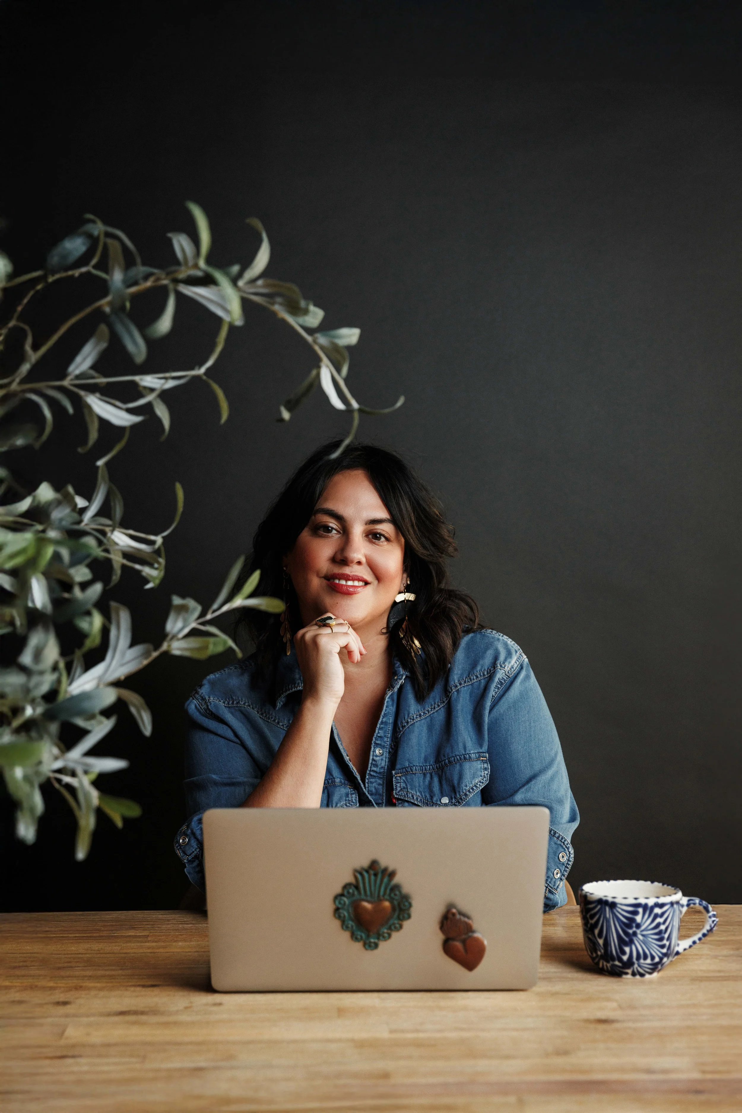 A woman with medium-length dark hair, wearing a denim shirt, sitting at a wooden table with a laptop adorned with decorative heart and paw stickers. She has a friendly expression, resting her chin on her hand, and is looking at the camera. To her right is a blue and white patterned ceramic mug, and to her left, there is a green plant with elongated leaves against a dark gray wall.