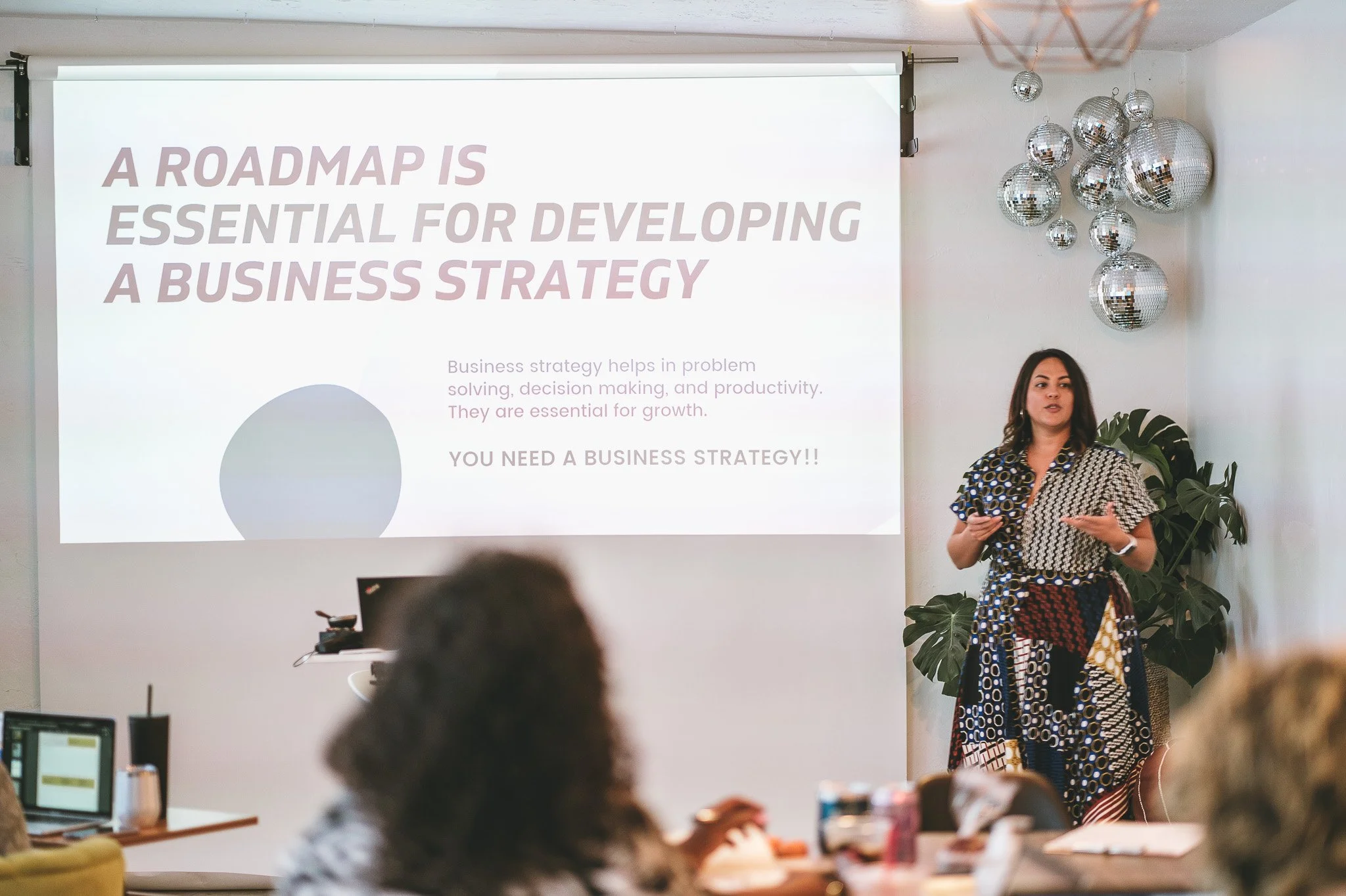A woman wearing a patterned dress is giving a presentation in a room decorated with disco balls and a plant. A slide on the screen reads: 'A ROADMAP IS ESSENTIAL FOR DEVELOPING A BUSINESS STRATEGY'. Several people are seated at tables, some with laptops and drinks, listening to the presentation.