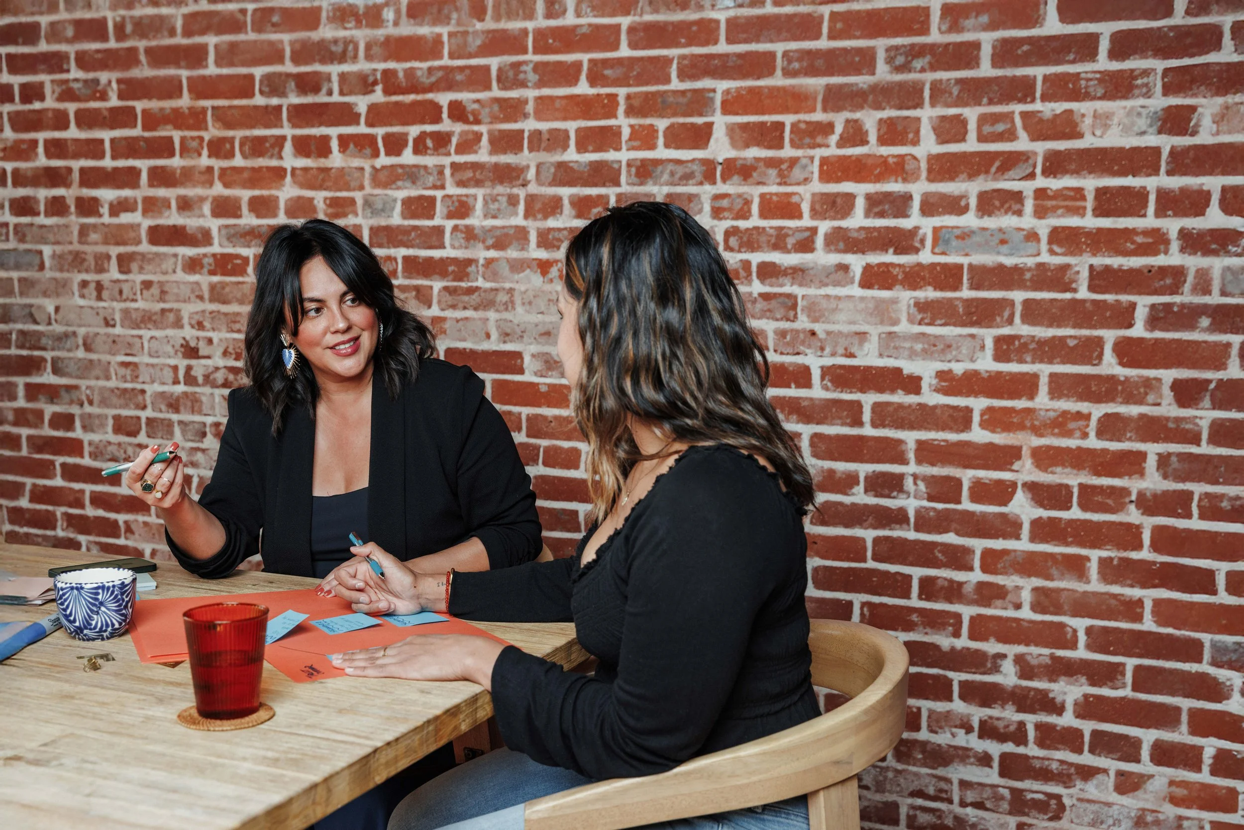 Two women engaged in conversation at a wooden table against a brick wall background. One woman has dark hair with waves, wearing a black top, and the other has dark brown hair with lighter highlights, also in black. The table has red paper, a blue and white mug, and red glasses.