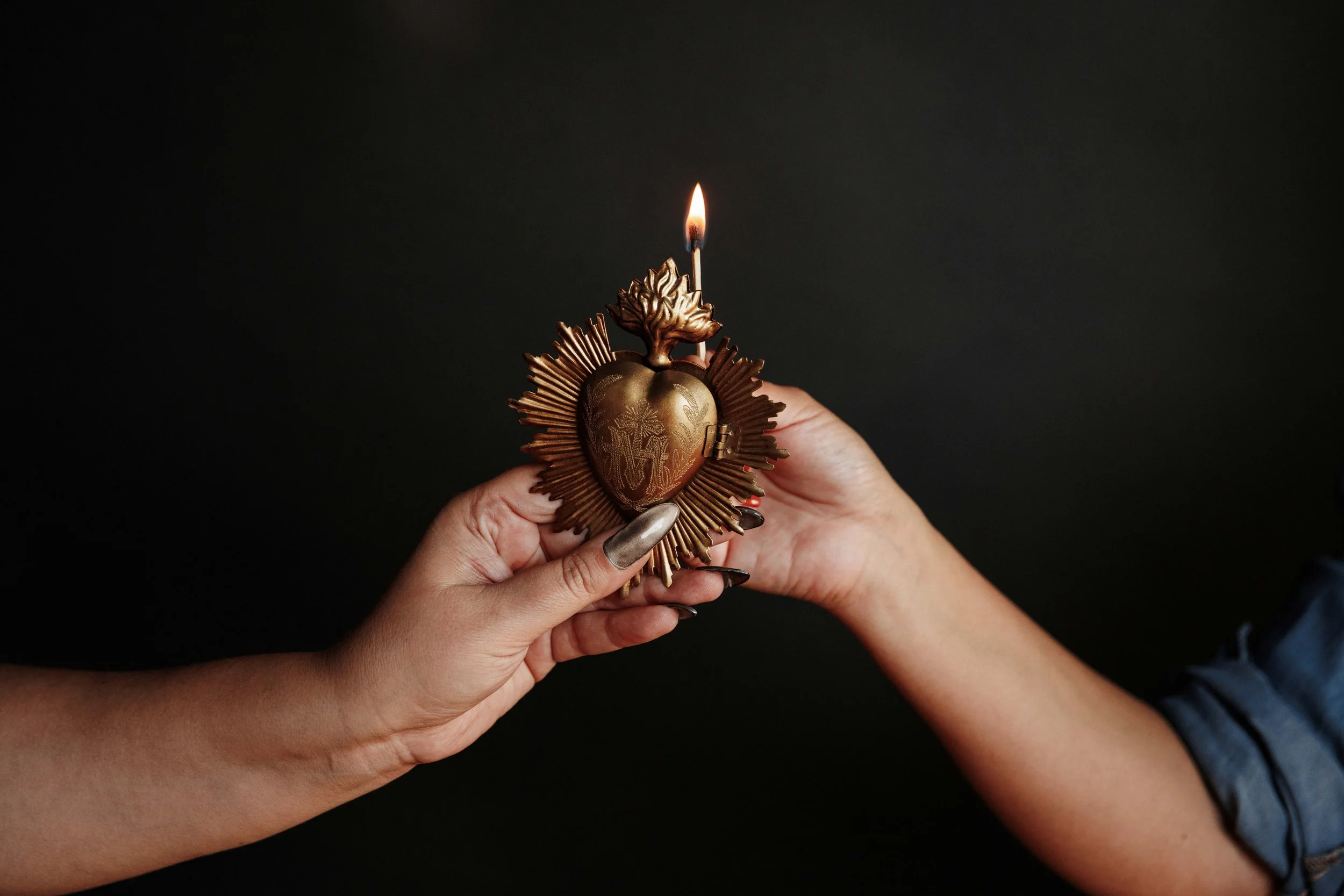 Two women's hands shown. One woman holiding a brass sacred heart. The other woman holding a match to light the top of the sacred heart with a flame.