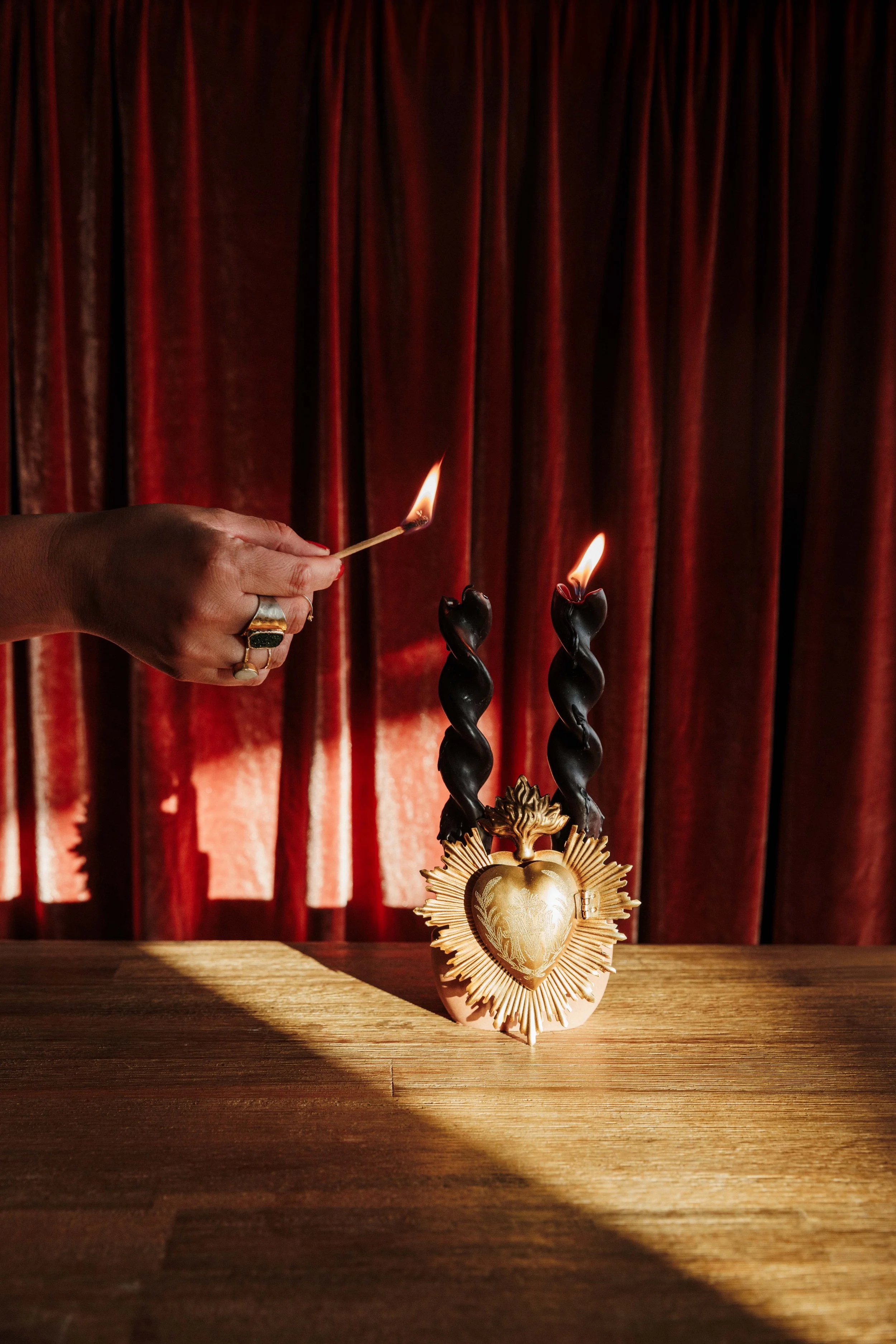 Woman hand with gold rings, lighting a black spiral candle. At the base of the candle is a brass sacred heart. The backgroun has terra cotta velvet curtains.