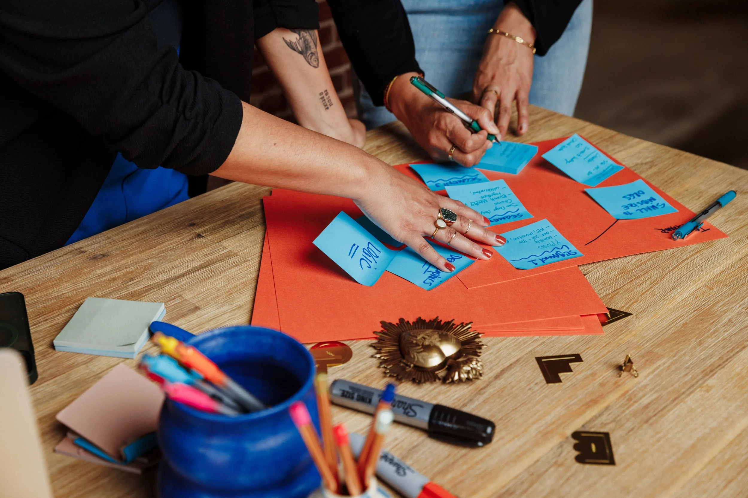 Picture of paper, a pen and pencil holder, with markers and sticky notes all over the table. Two sets of hands are organizing the sticky notes.