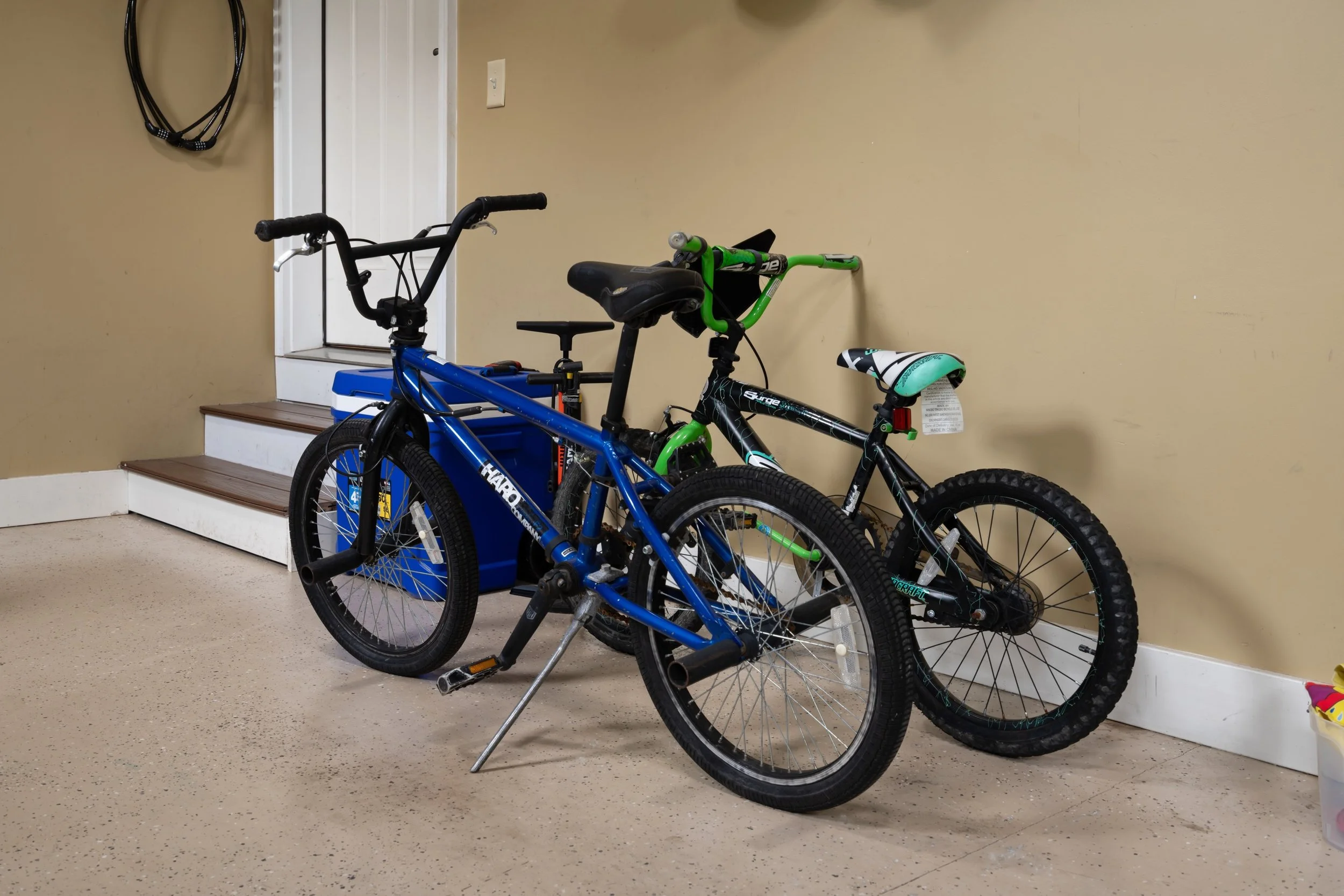 Two bicycles, one blue and one black and green, leaning against a beige wall near a set of stairs. There is a small yellow container with a pink handle on the right side of the image.