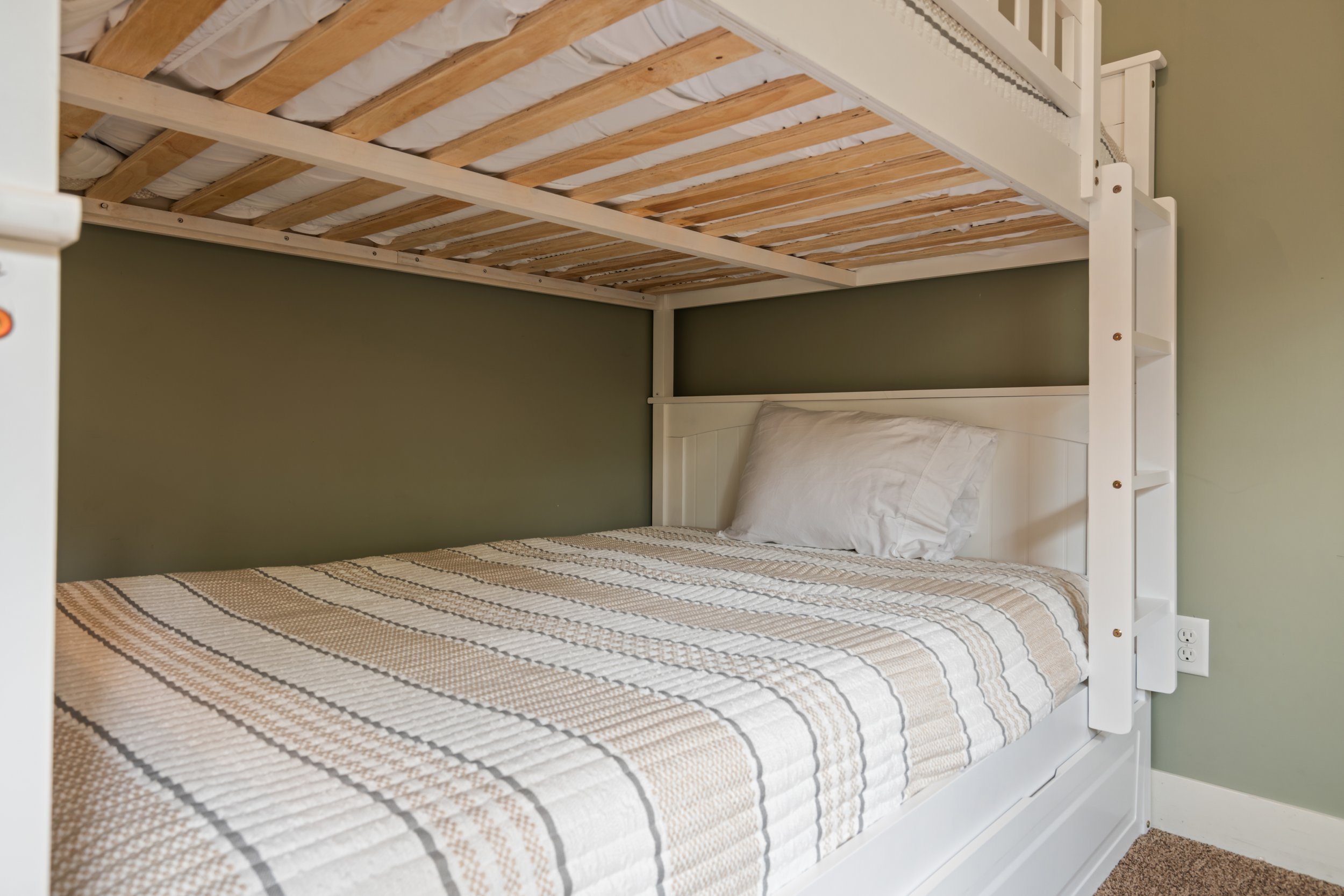 A bedroom with a bunk bed featuring a white frame and striped bedding in beige, brown, and white. The lower bunk has a white pillow and a small section of carpet is visible on the floor.