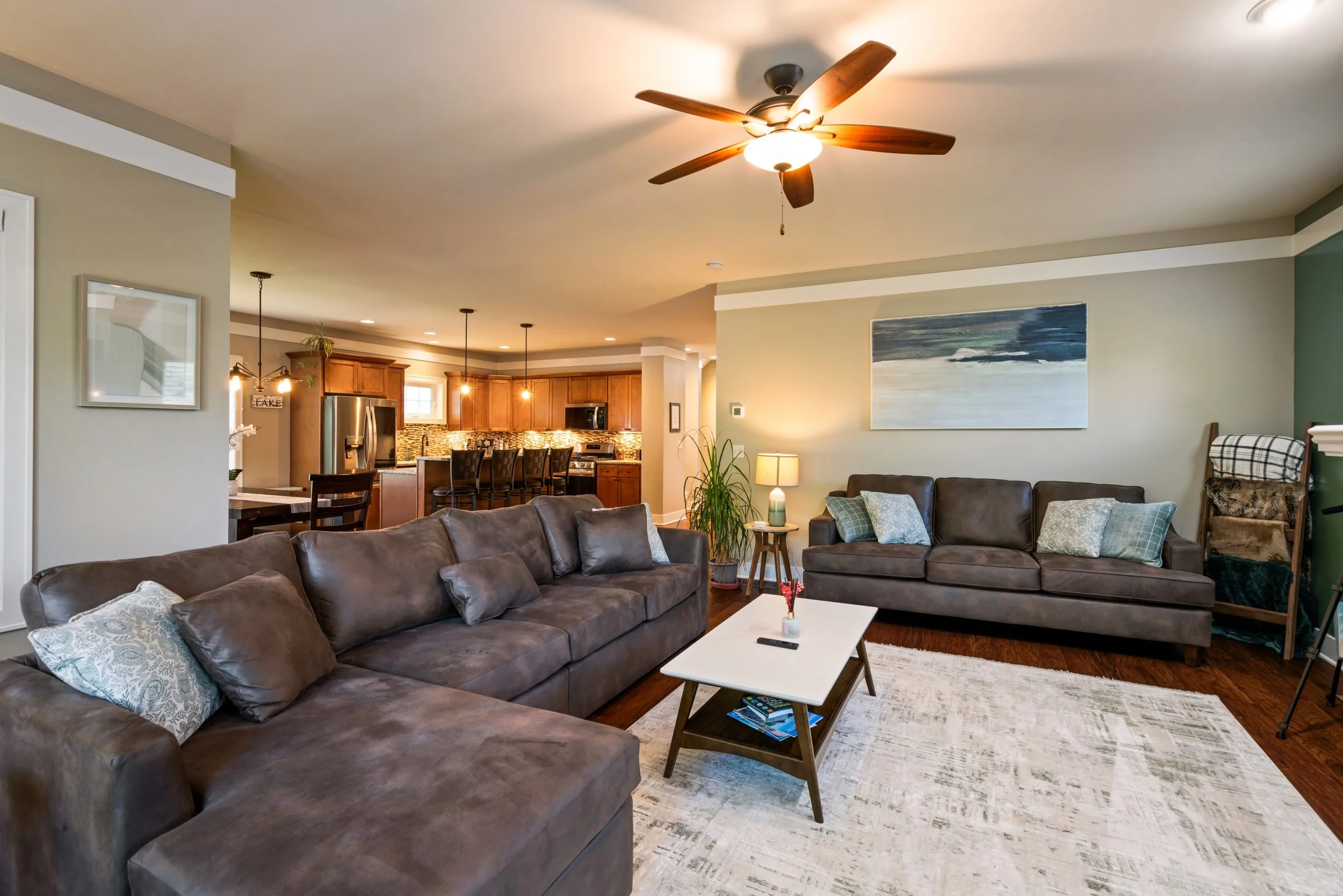 Living room with brown leather sofas, a white coffee table, framed artwork on light green walls, a ceiling fan, and an open kitchen area with wooden cabinets and pendant lights.