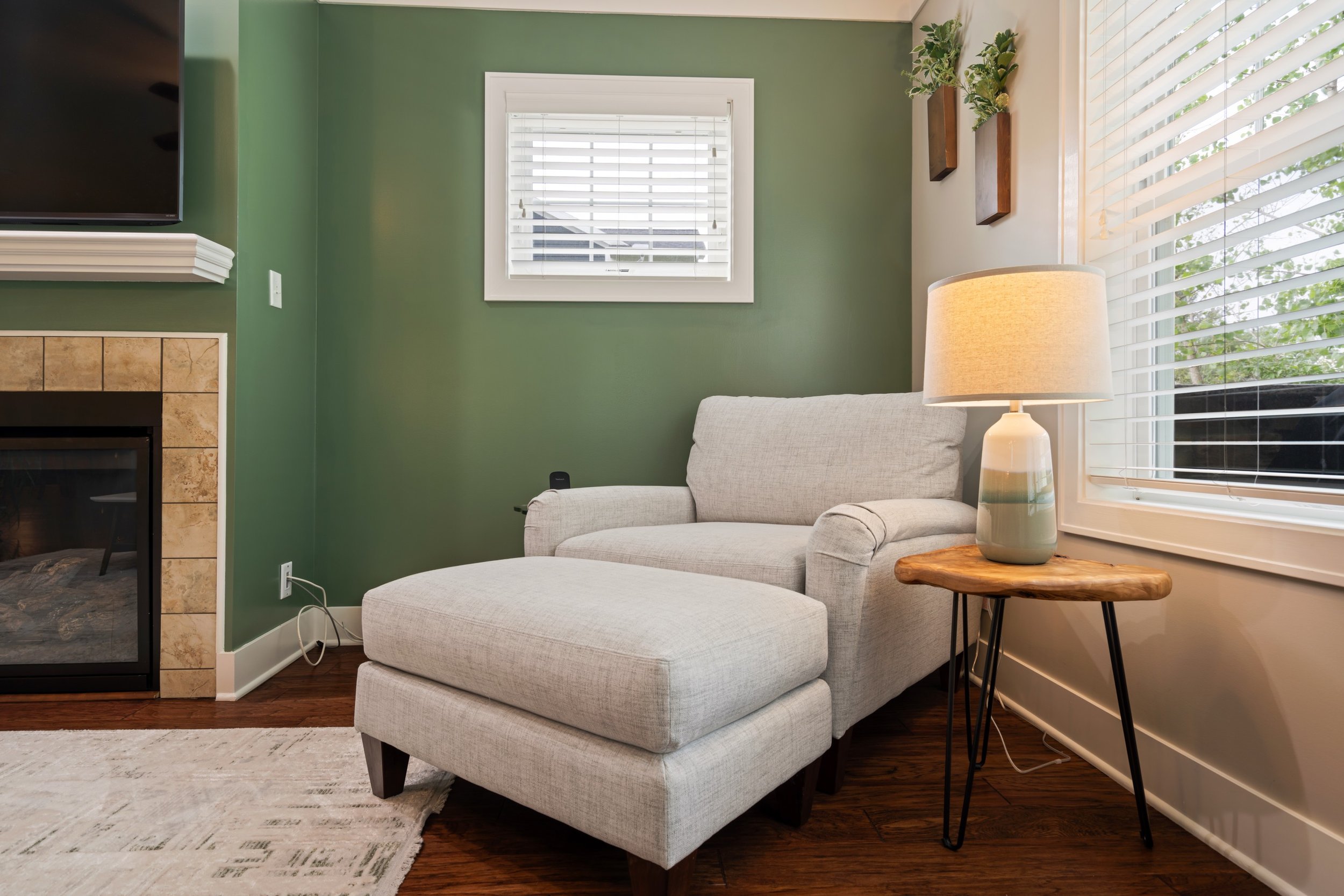 A cozy living room corner with a beige armchair, a wooden side table with a lamp, a window with white blinds, and green walls decorated with small potted plants on wooden shelves.