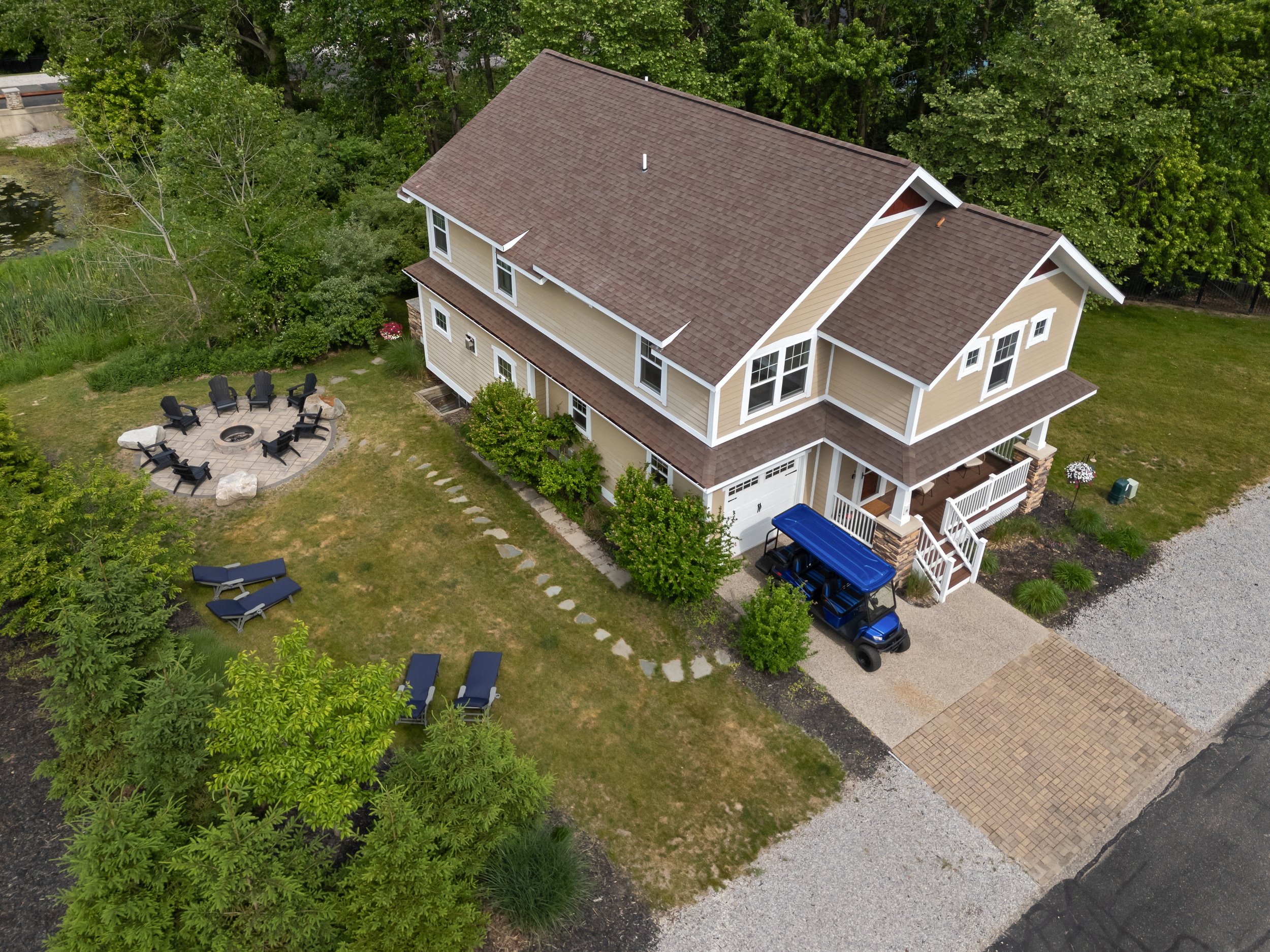 Aerial view of a beige, two-story house with a brown shingle roof, surrounded by lush green trees, a gravel driveway, and outdoor seating with chairs around a firepit.