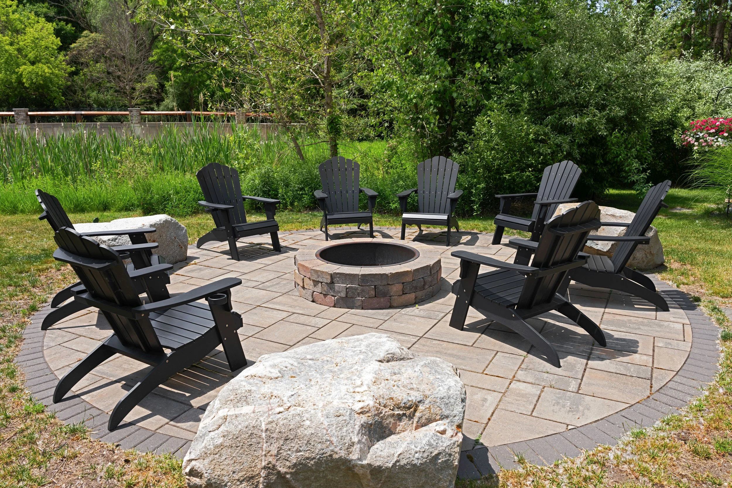 A backyard sitting area with eight black Adirondack chairs arranged in a circle around a brick fire pit on a paved patio. Large rocks surround the patio, with green grass and lush shrubs and trees in the background.