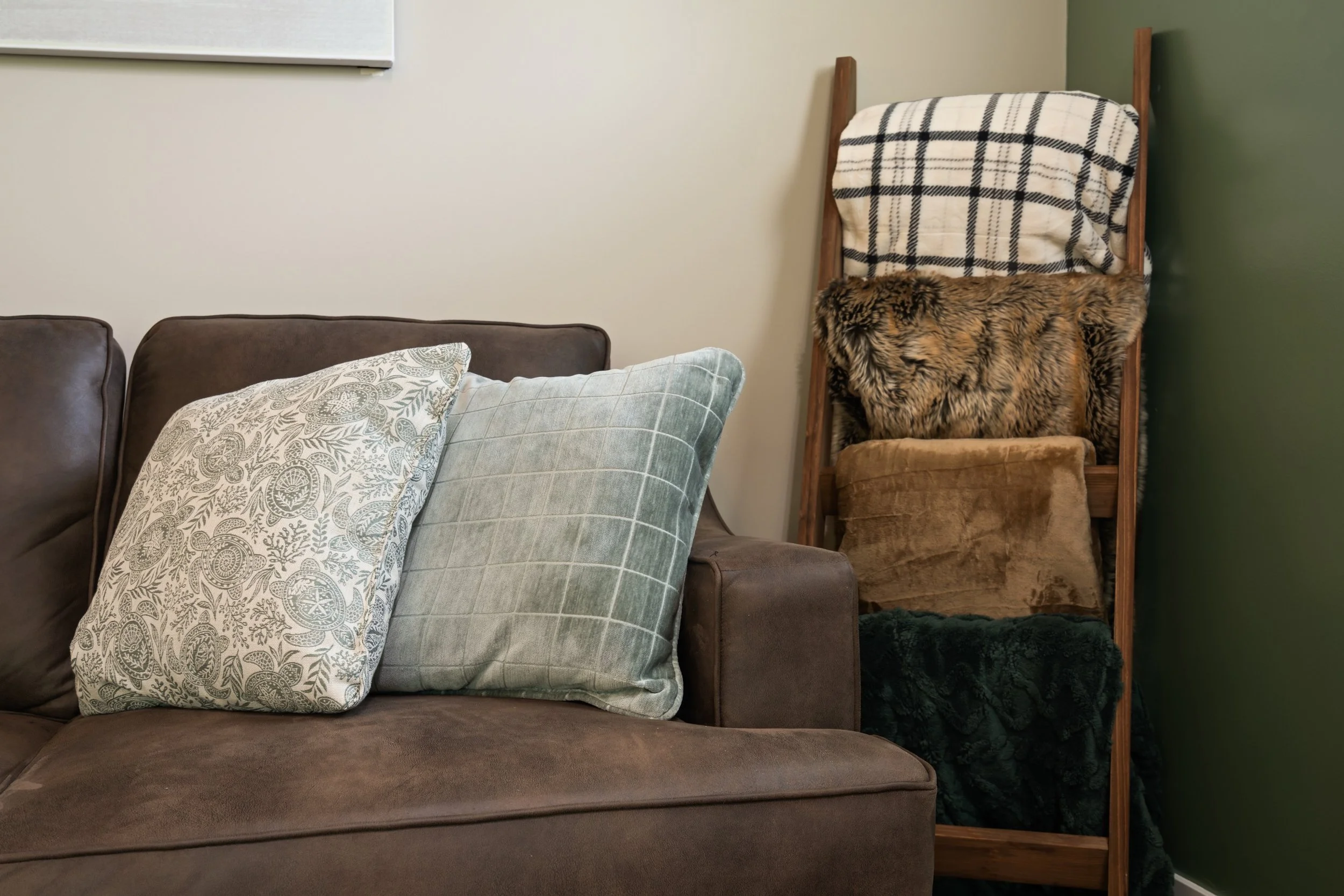 A brown couch with two decorative pillows, one with a white floral pattern and the other with a blue plaid design. There is a wooden ladder in the background with a plaid blanket and fur covering on the upper part, against a green and beige wall.