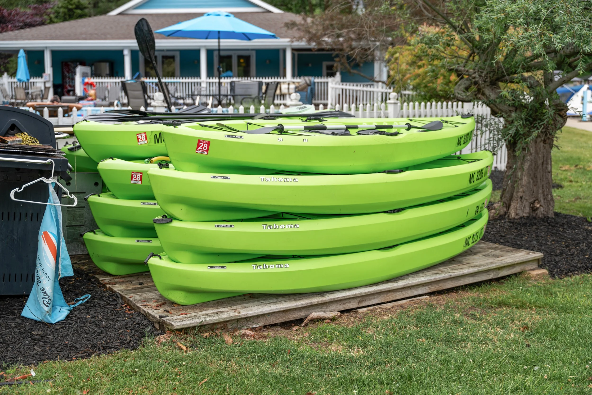 A stack of green kayaks labeled 'Tahoe' and with registration number 'MC 8206 UE' is placed on a wooden platform beside a lush green lawn and a tree.