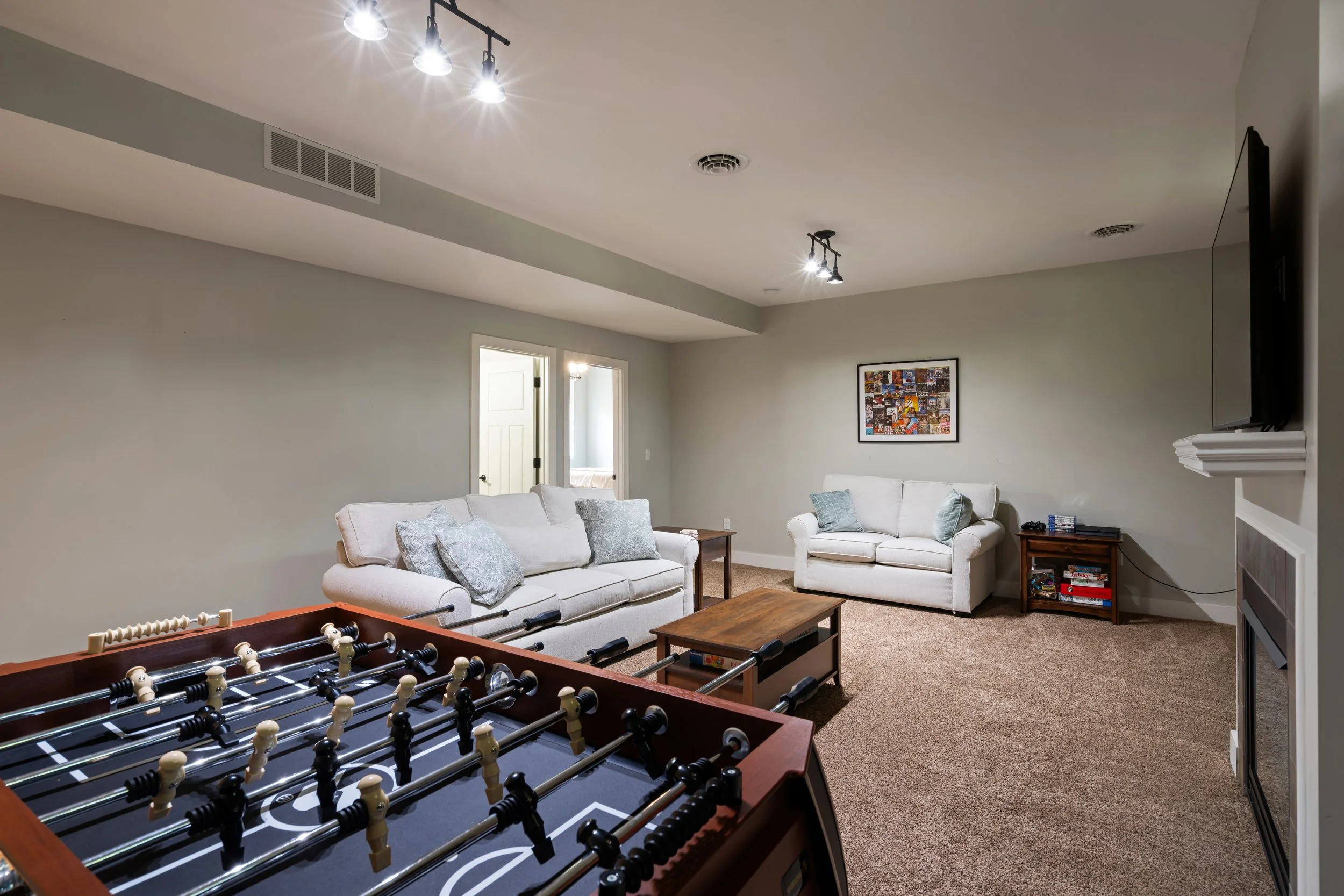 Living room with three beige sofas, gray pillows, a wooden coffee table, a framed collage on the wall, a black TV mounted on the wall, and a foosball table in the foreground. Ceiling lights are turned on, with a beige carpet and doors leading to other rooms.