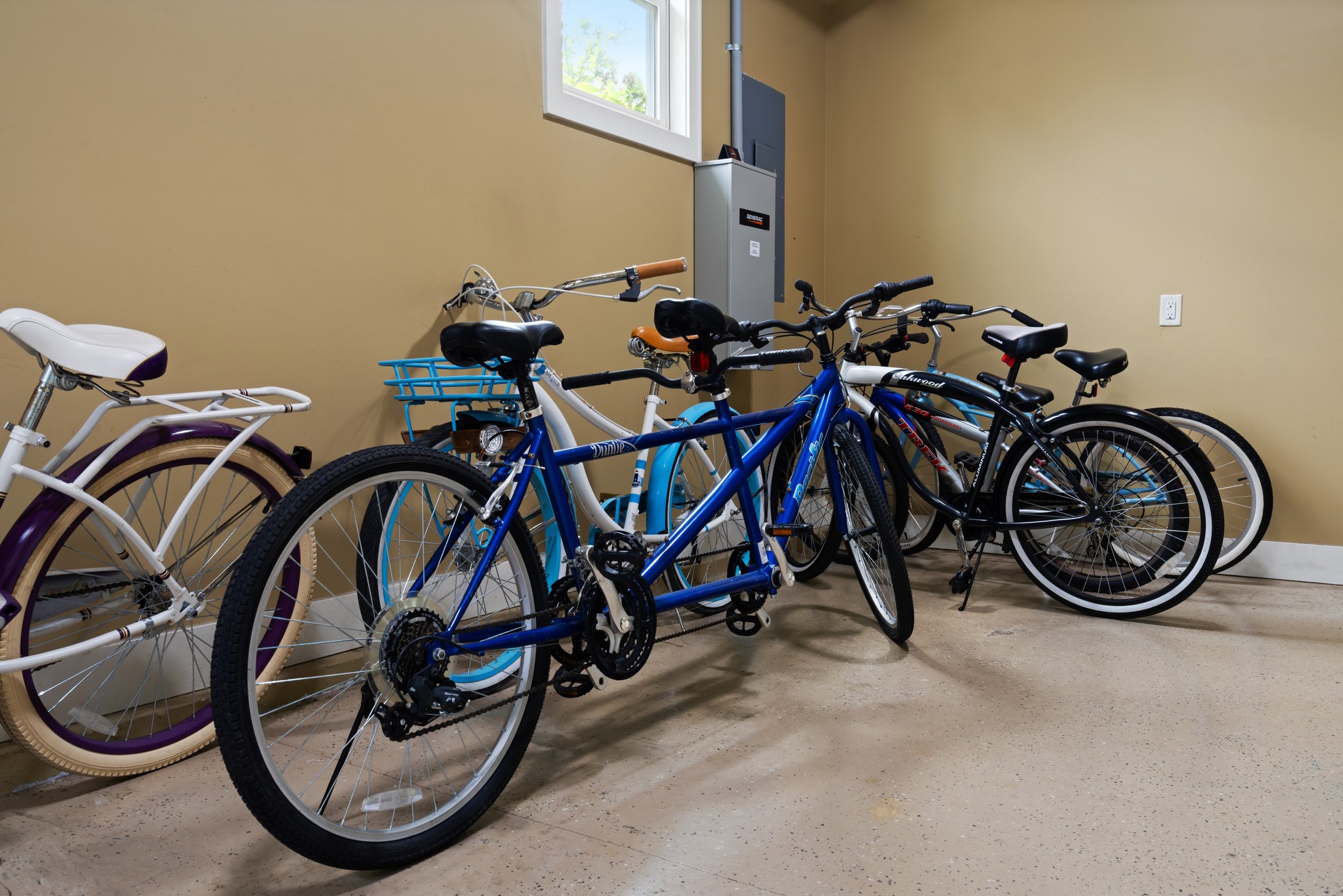 A room with pale yellow walls and a window, containing several bicycles including a white bike with purple accents, a blue bike, and black bikes lined up next to each other.