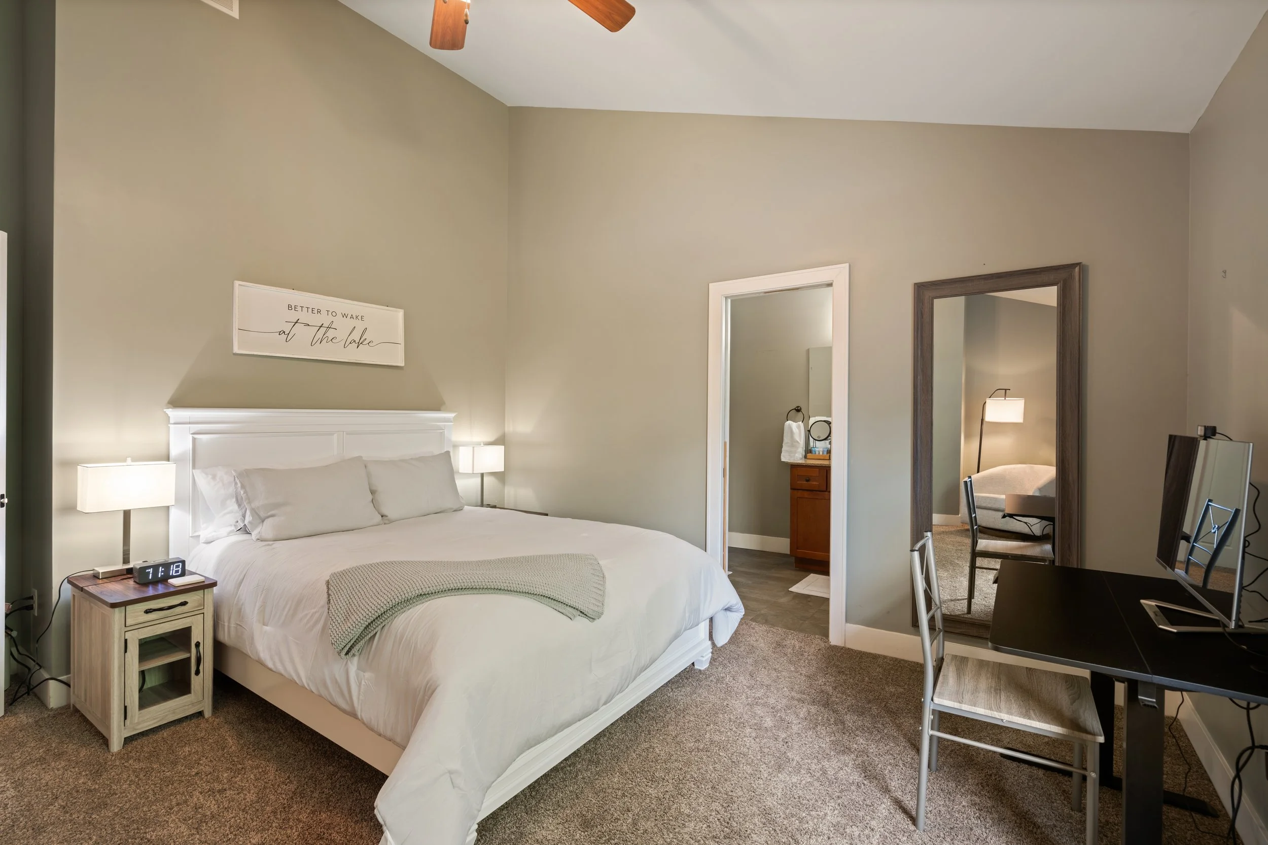 A bedroom with a white bed, nightstand, and lamp, beige walls, and a window. There is a mirror and a doorway leading to a bathroom, with a chair and a side table visible in the background.