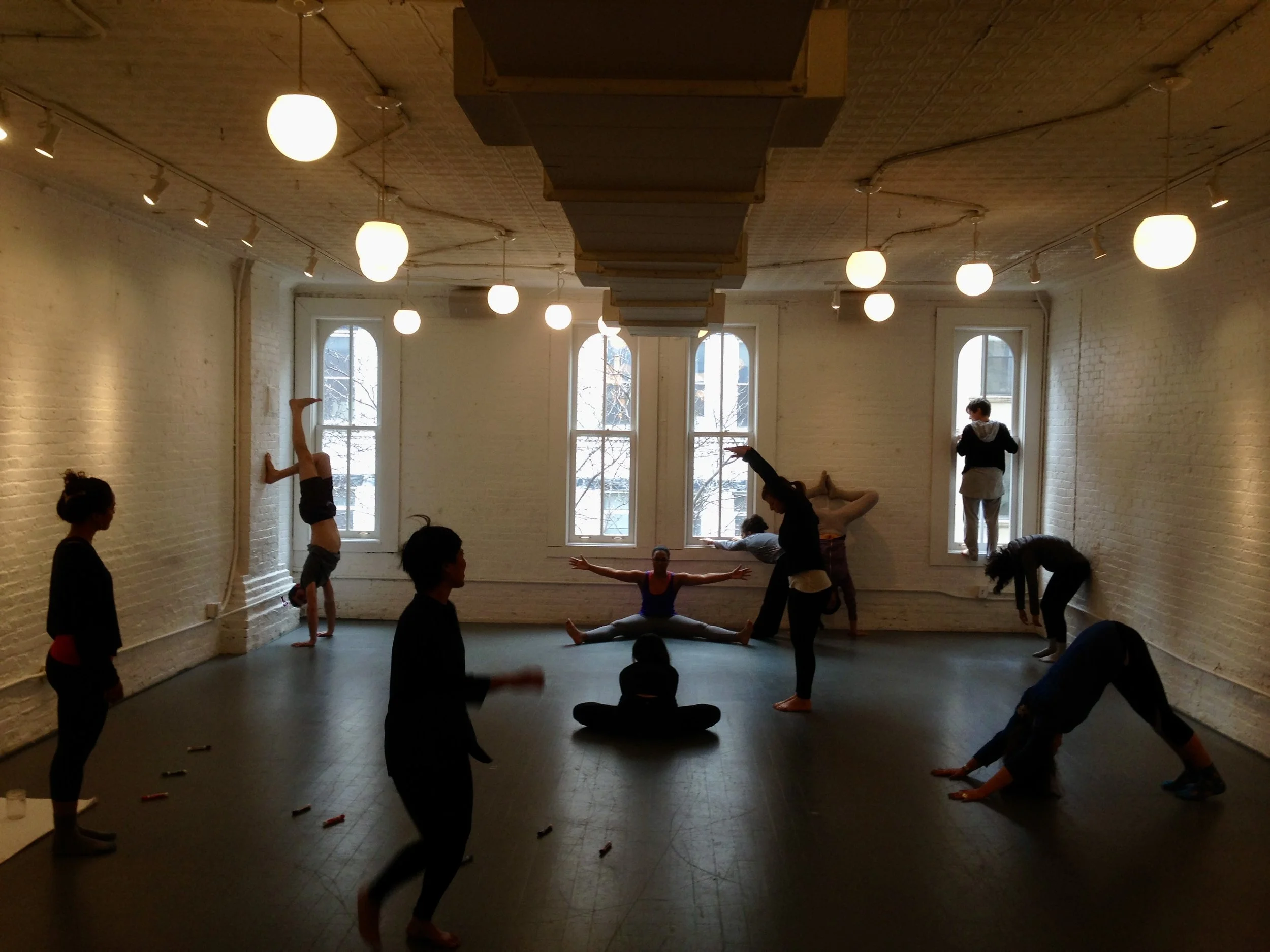 A group of people practicing yoga or stretching exercises in a spacious room with white brick walls and large windows, with some individuals doing handstands, splits, and backbends.